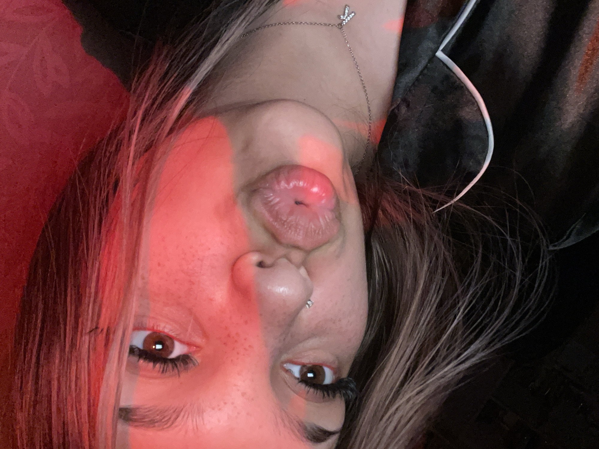 A close-up, angled selfie shows a young Caucasian female with brown eyes, brown hair with blonde highlights, and fair skin with freckles. She is wearing a silver necklace with a letter "K" pendant. She has her lips pursed, and there is a piercing on her lower lip. She is wearing a black garment with white trim. The lighting in the image is predominantly red, casting a red hue over her face and hair. The background is dark and appears to be indoors. There are no visible indicators of a specific city or country, including Unknown, Russia.