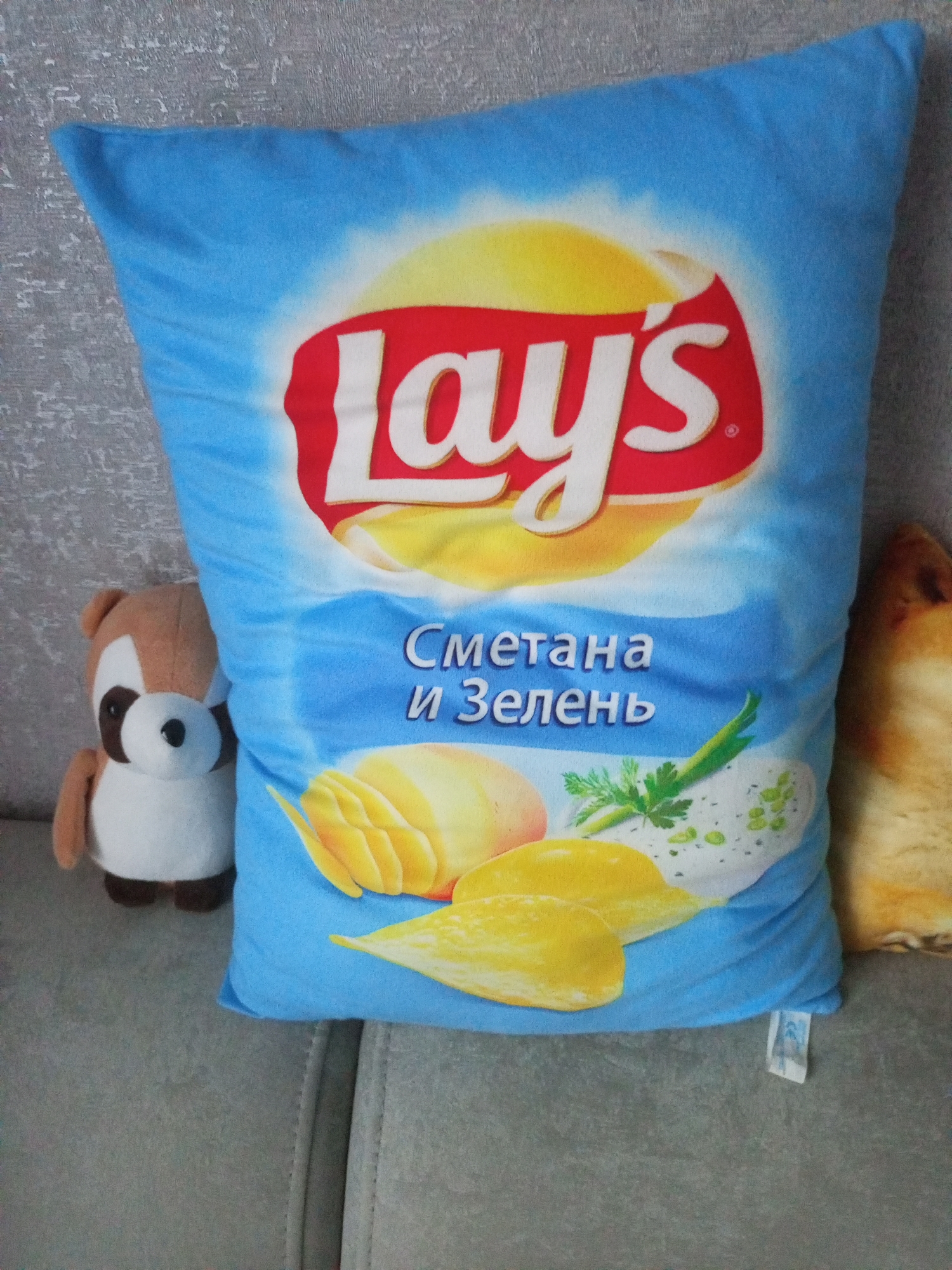 A close-up, eye-level shot shows a light blue pillow designed to look like a bag of Lay's potato chips. The pillow is situated on a grey sofa. To the left of the pillow, a stuffed toy resembling a squirrel with brown and white fur is visible. To the right, the fuzzy ear and side of what appears to be a dog are partially visible. The pillow is the main focus, displaying the Lay's logo in red and white, a yellow sunburst, and images of potato chips with a creamy dip and green herbs. Text in Cyrillic script, reading "Сметана и Зелень" (Sour cream and greens), is present on the pillow. The background is a textured grey surface, possibly a wall. The scene is indoor, and no specific landmarks or elements place it within the city of Homyel, Belarus.