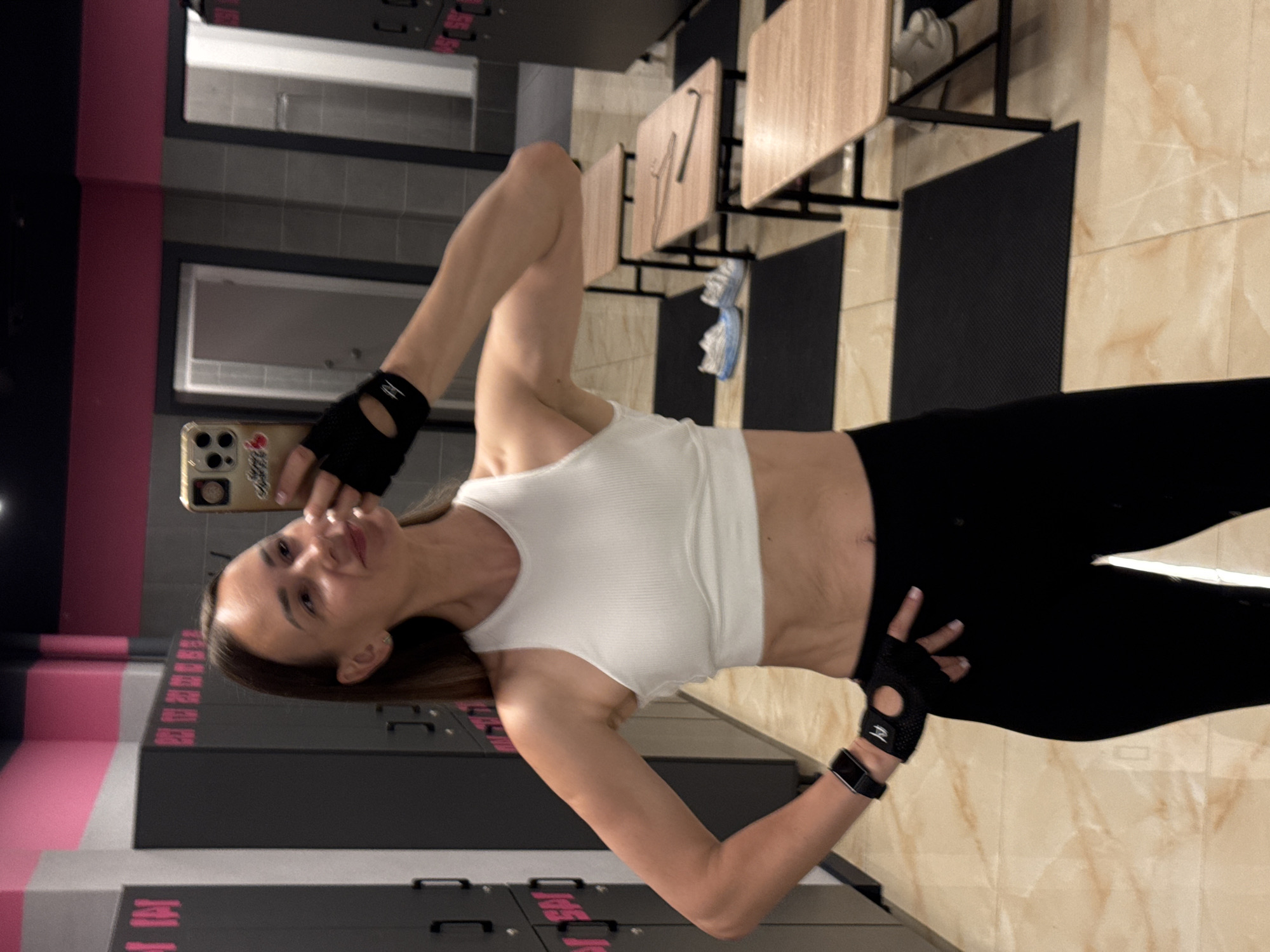 A woman is taking a selfie in a gym locker room. She is wearing a white tank top, black leggings, black workout gloves, and a black smartwatch. She is smiling and has her right arm raised. In the background, there are dark gray lockers with pink trim, wooden benches, and black mats on the floor. The room appears to be clean and well-lit. The location is the city of Sochi, Russia.
