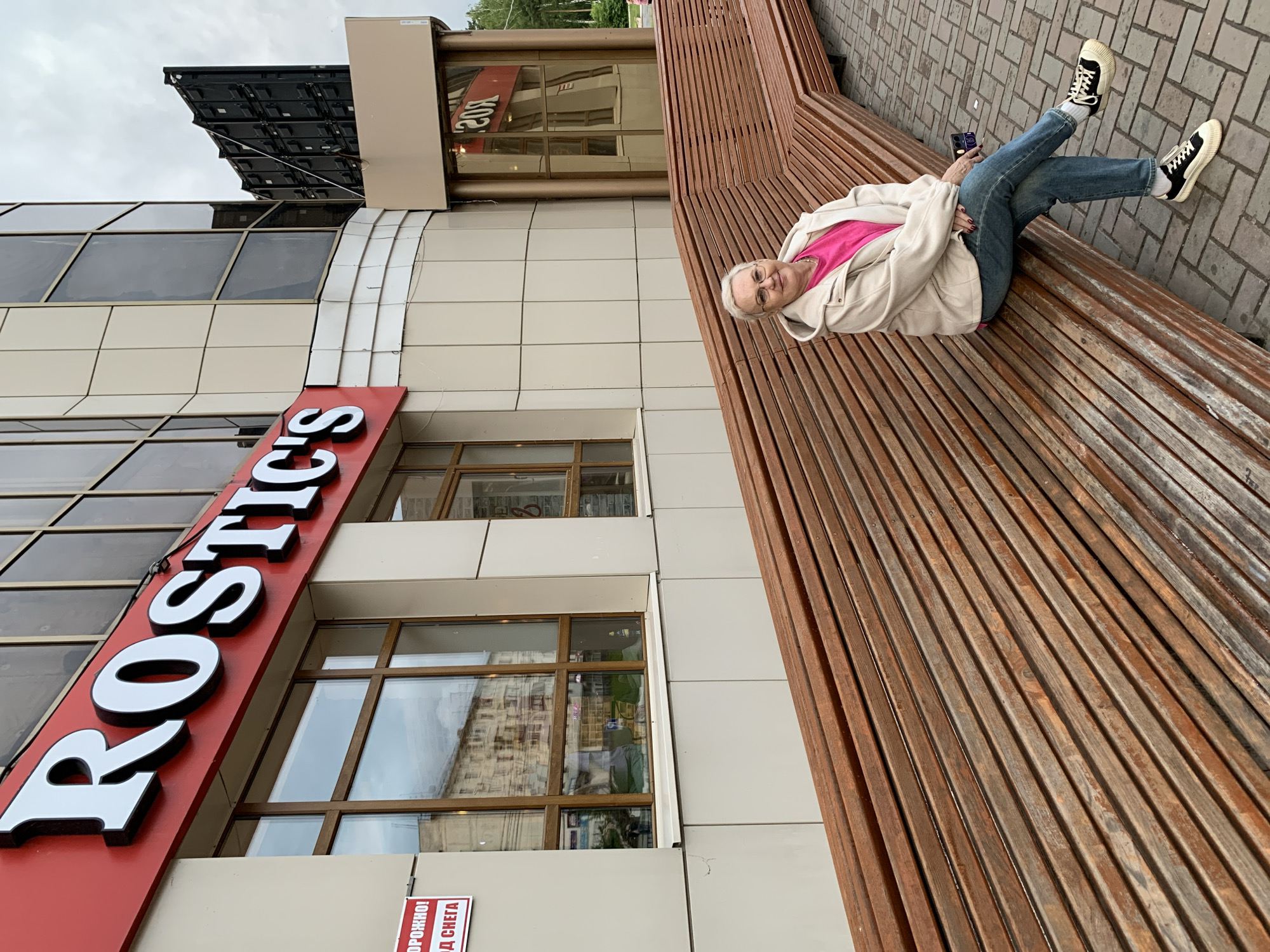 An elderly woman with white hair and glasses is seated on a long, multi-angled wooden slatted bench in the city of Chaikovsky, Russia. She is wearing a bright pink top, a light beige hooded jacket, blue jeans, and black and white sneakers. Her legs are crossed, and she is holding a dark-colored smartphone in her left hand. The woman is looking directly at the camera with a slight smile.
Behind her is a modern multi-story building with light-colored paneling and large rectangular windows, which reflect a residential building and the overcast sky. A prominent red sign with white, black-outlined letters spelling "Rostic's" is mounted above the ground-floor windows of the building. To the left of the Rostic's sign, a red and white warning sign with partially visible Cyrillic text, reading "ОРОЖНО!" (DANGER!) and "Д СНЕГА" (OF SNOW), is affixed to the wall. To the right of the building, a large, dark, modular outdoor LED screen or billboard structure is visible against the overcast sky, with green foliage from distant trees appearing further right. The foreground consists of light brown and grey brick-patterned paving stones.