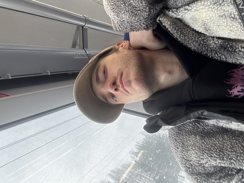 A young adult male, likely Caucasian, is depicted in a close-up selfie taken inside a public transport vehicle, presumably in the city of Redkino, Russia. He is facing the camera with a direct gaze, wearing a beige baseball cap, a black hooded sweatshirt with a partially visible purple graphic, and a mottled gray and white sherpa fleece jacket, partially unzipped. His right hand is positioned behind his neck. The interior of the vehicle features a light-colored ceiling with horizontal panels, a vertical silver support pole on the right, and a horizontal white bar with the alphanumeric inscription "474.48.SD". Through the large window occupying the left and upper portions of the frame, a winter landscape is visible, characterized by snow-covered trees and an overcast sky. Overhead wires, possibly power lines, traverse the exterior view.