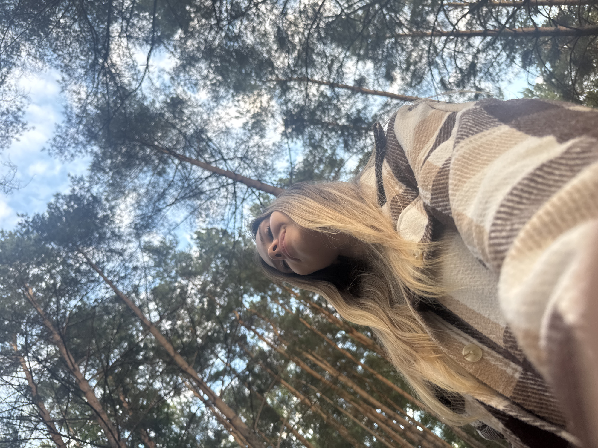 A young woman with blonde hair is captured from a low angle, looking up at the sky. She is wearing a plaid shirt with brown and tan colors. Her eyes are closed, and she has a slight smile on her face, suggesting she is enjoying the moment. The background consists of tall pine trees with a blue sky and some white clouds visible through their branches. The image appears to be taken outdoors in a wooded area, possibly a park or forest. Without further context, it is not possible to pinpoint the exact location within the city of Chaikovsky, Russia.