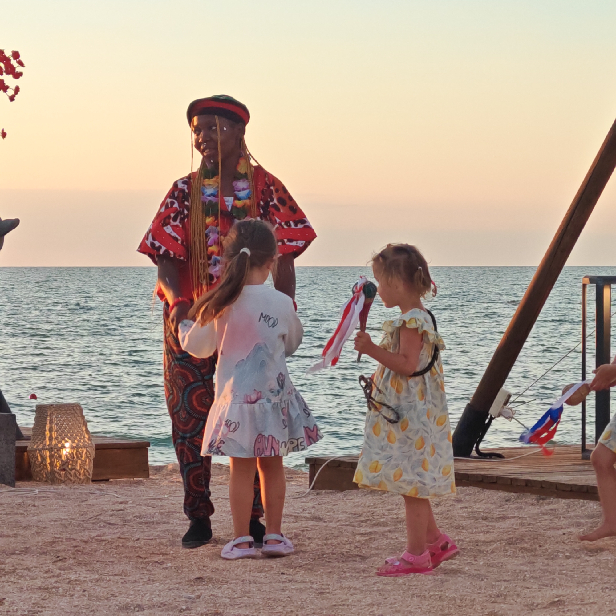 A group of people are gathered on a sandy beach near the sea at sunset. The main subjects are a performer in colorful traditional attire and two young girls. The performer, a dark-skinned individual wearing a red and black patterned top, a beaded necklace, and a distinctive hat, appears to be interacting with the children. The two girls are positioned in front of the performer. One girl, wearing a white dress with a pastel mountain and floral print, stands facing away from the camera. The other girl, in a yellow dress adorned with lemon patterns, faces the performer and holds a stick with red and white ribbons. The background features a body of water, likely the sea, with gentle waves. The sky is a soft gradient of orange and yellow, indicative of a sunset. On the left side of the image, a woven basket with a light inside is visible, along with part of a wooden structure. On the right, a wooden pier or deck is present, with a rustic wooden pole. The overall scene suggests an outdoor event or performance taking place by the coast. The specific location is described as the city of Shchyolkine, Ukraine.