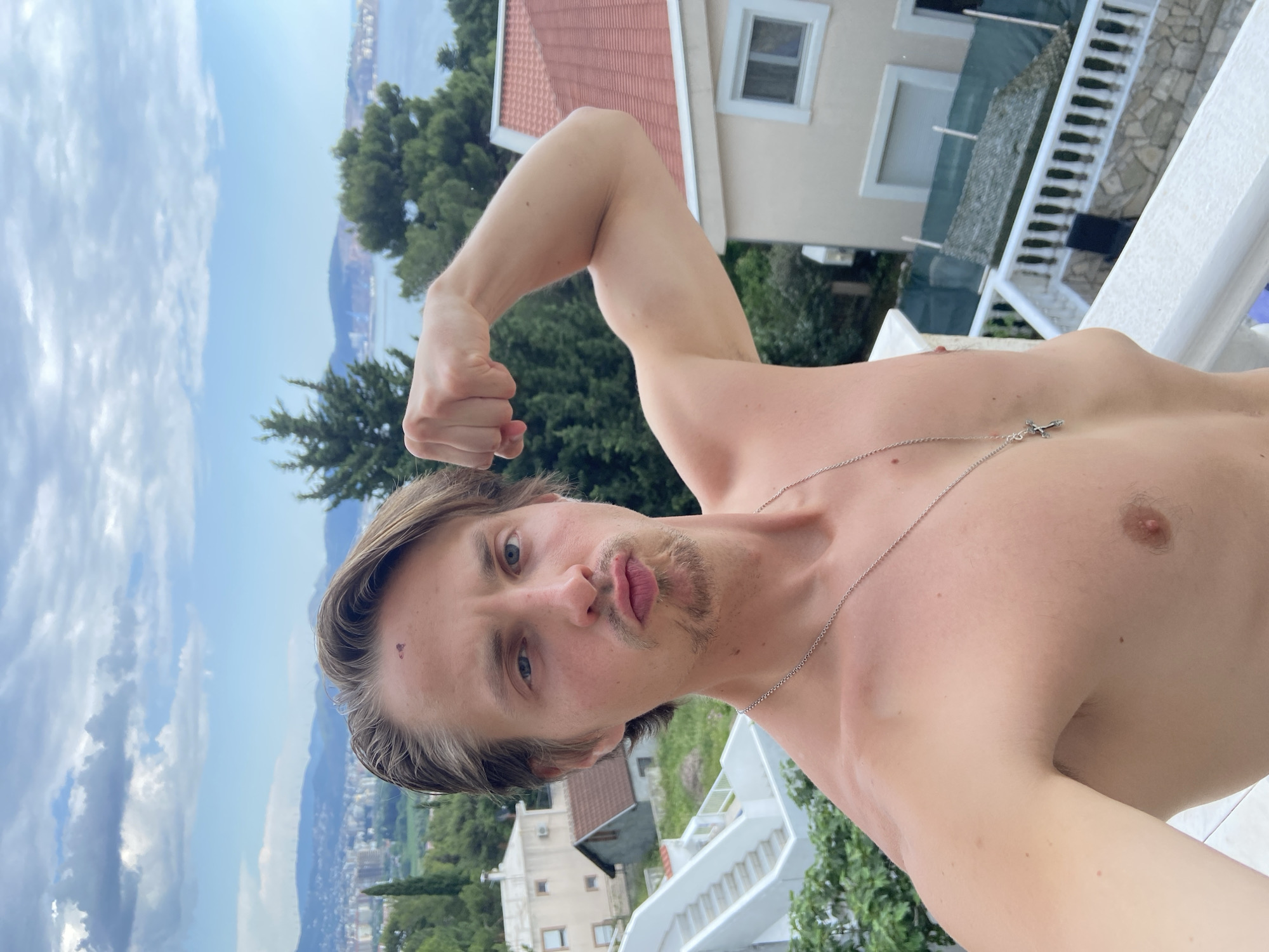 A shirtless man with fair skin, light brown hair, and blue eyes is posing for a selfie in Šušanj, Montenegro. He is flexing his right bicep, and is wearing a silver necklace with a cross pendant. The background features the sea and sky, buildings, and greenery characteristic of the location. Specifically, some buildings have red-tiled roofs. The sky is partly cloudy. The man appears to be on a balcony or terrace with white railings.