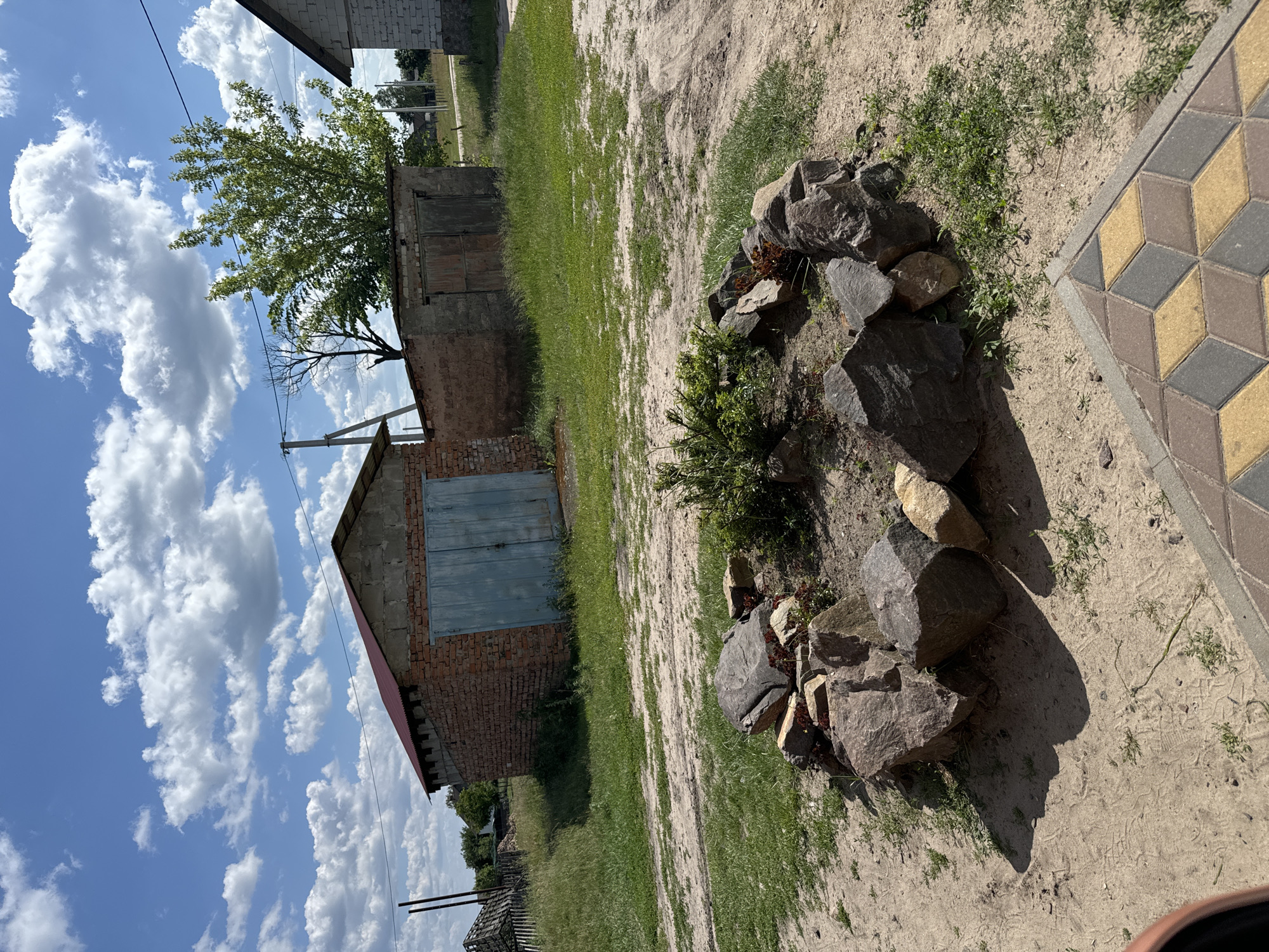 This is a photograph of an outdoor scene with a brick building, a bonfire pit, and a paved area. The building is made of red brick and has a large, light blue garage door. A tree with green foliage is growing next to the building. The bonfire pit is made of large rocks and is situated on sandy ground with patches of grass. A section of paved ground with a geometric pattern of yellow, grey, and brown tiles is visible on the right side of the image. The sky is blue with large, white clouds. The lighting suggests it is daytime. The image is taken from a slightly elevated, tilted perspective. There is no text visible in the image.