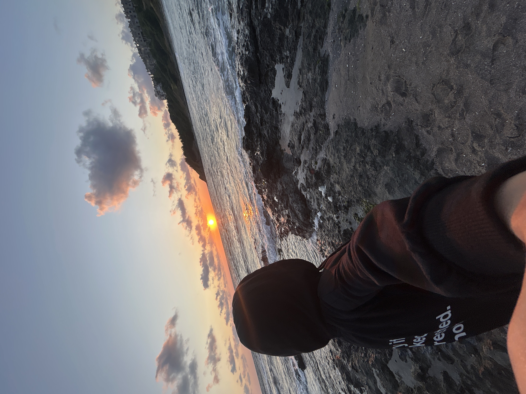 A person wearing a black hooded sweatshirt with white text on the back stands on a rocky beach, facing the ocean at sunset. The sun is low on the horizon, casting a warm glow over the water and the sky. The waves are gently crashing against the shore. In the background, a line of buildings can be seen on a hillside. The person's hand is outstretched, suggesting they are taking a selfie. The text on the sweatshirt is partially visible and appears to say "try it, beeker, rested. no". The overall scene depicts a serene and picturesque moment at the coast of Tsarevo, Bulgaria.