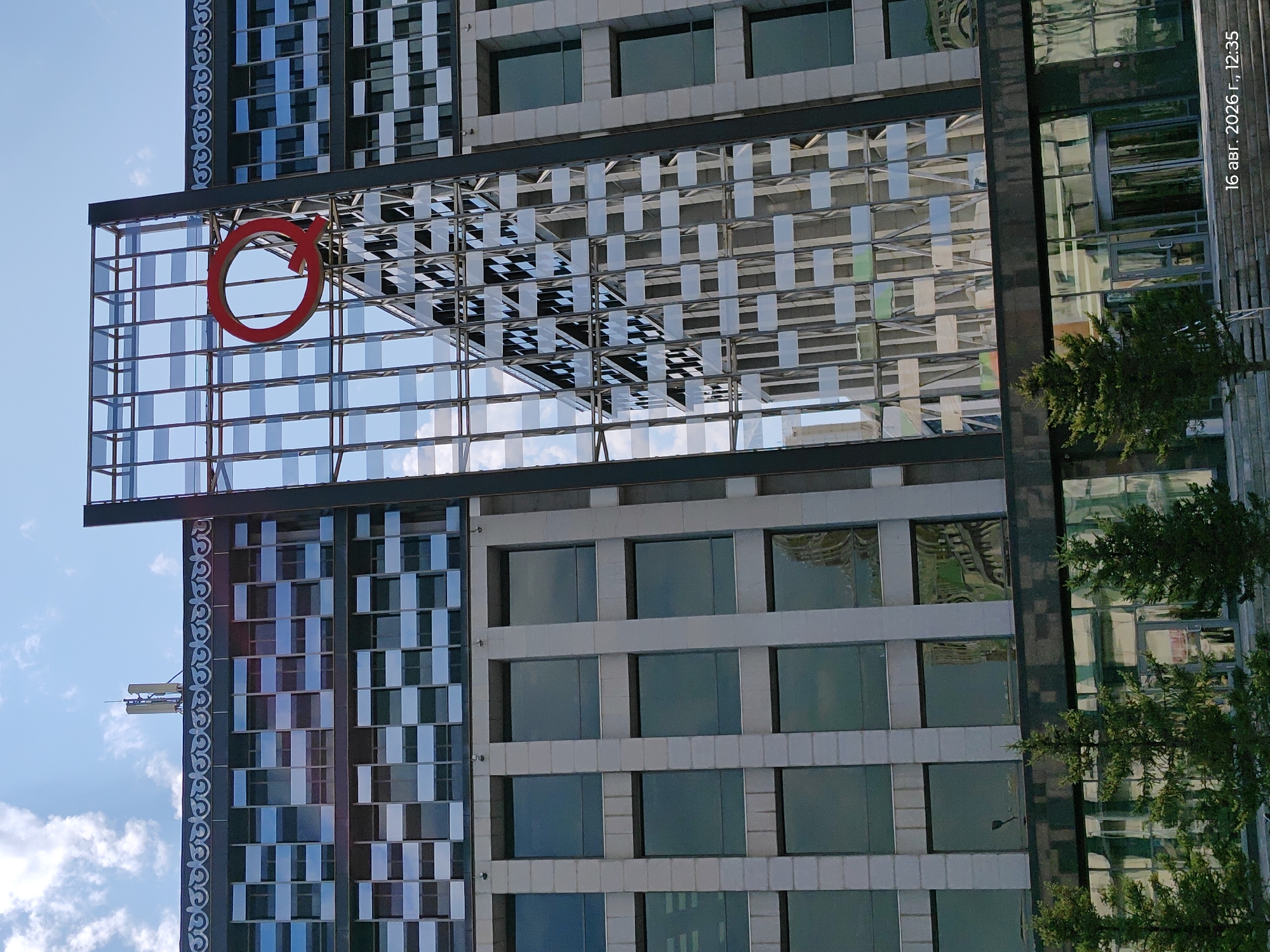 This is a medium-angle, full-shot photograph of a modern, multi-story building in Astana, Kazakhstan. The building features a predominantly glass facade with contrasting elements of light-colored stone and dark metal. A prominent red circular logo, resembling the letter "Q," is affixed to the glass structure on the right side of the building. The sky above is a clear blue with scattered white clouds. In the foreground, several evergreen trees are visible, and the lower part of the building has a glass entrance. A timestamp in the bottom right corner reads "16 авг. 2026 г., 12:35" which translates to "August 16, 2026, 12:35". The overall scene depicts a contemporary urban architectural design.