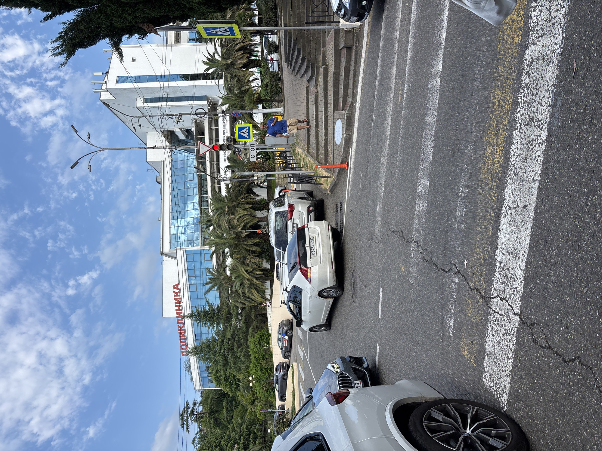 A street scene in Sochi, Russia, depicts a building with "ПОЛИКЛИНИКА" (Clinic) written in red letters above a facade of blue glass windows. Several cars are stopped at a traffic light, with a pedestrian crossing visible in the foreground. A man in a blue t-shirt and shorts walks down a set of outdoor stairs. Palm trees and other greenery surround the area, under a partly cloudy sky.