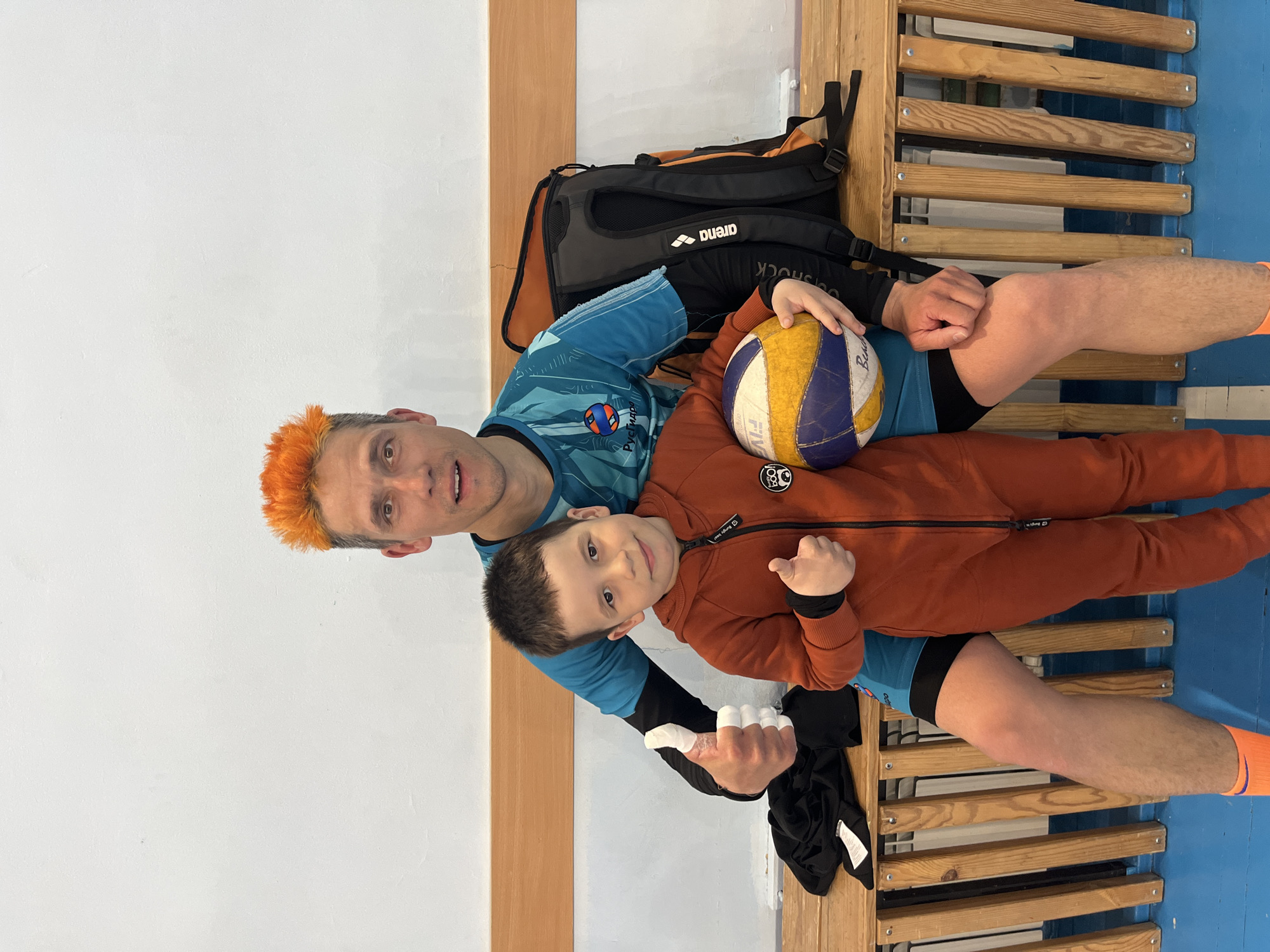 Two individuals, an adult and a child, are seated on a wooden slat bench. The adult has short hair dyed orange and is wearing a blue and black sports jersey and orange socks. The child is wearing an orange zip-up hoodie with black sleeves. The adult is holding a volleyball. A black and orange backpack is resting against the wall behind them. The adult has tape on his fingers. The location is in Zav'yalovo, Russia. The room has a white and tan wall. The floor is blue.