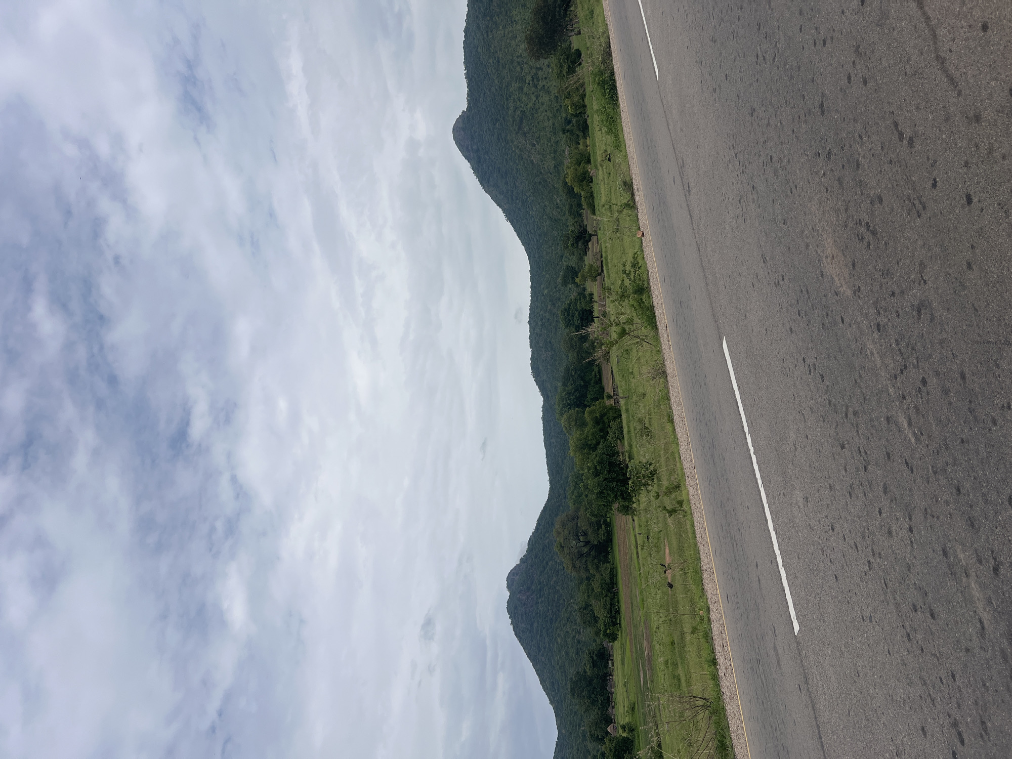 The image depicts a scenic view of a rural landscape in Minda, Nigeria, characterized by rolling green hills covered in dense vegetation and scattered trees. A paved road with a white dashed line runs diagonally across the foreground, suggesting it is a route through this region. The sky above is overcast with a layer of clouds. In the mid-ground, the terrain features open grassy fields and more clusters of trees. A few small structures, possibly huts or houses, are visible nestled amongst the trees on the left side of the image. The overall impression is one of natural beauty and rural tranquility.