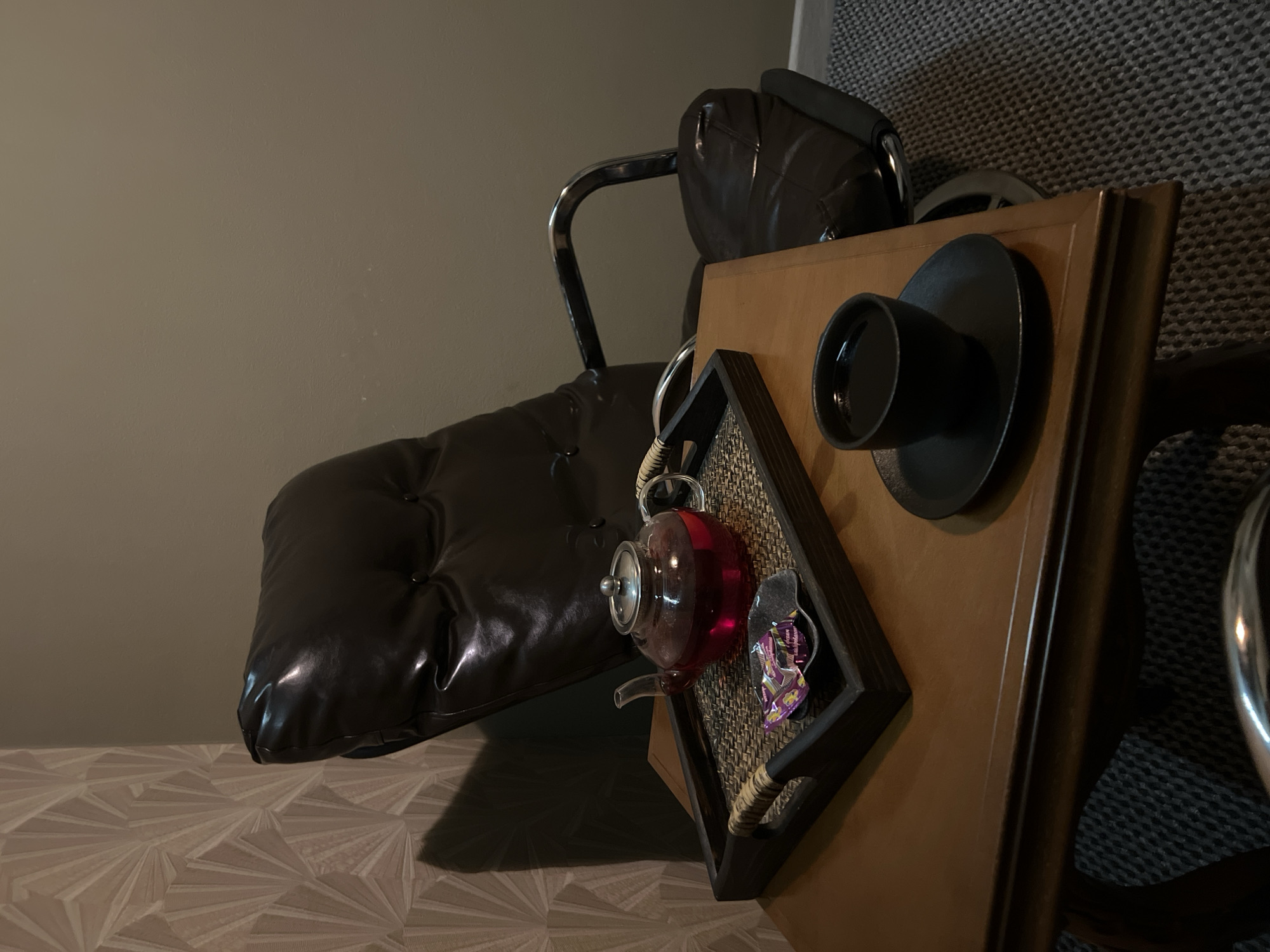 The image depicts an indoor scene in what is stated to be the city of Tyumen, Russia. In the foreground, a dark wooden side table is positioned to the left of a dark brown or black upholstered armchair with a high back and polished metal frame and armrests.
On the table, a dark wooden serving tray with woven rattan-style handles holds a clear glass teapot containing a translucent, deep red liquid. Adjacent to the teapot on the tray is a small, dark, irregularly shaped dish containing two individually wrapped candies, primarily purple and gold in color. To the right side of the table, a black ceramic cup filled with a dark liquid rests on a matching black ceramic saucer.
The armchair is situated against two distinct walls. The wall to the left features patterned wallpaper with a repeating beige or light brown geometric design, reminiscent of fanning motifs. The wall to the right is painted a plain, muted taupe or beige color. A distinct shadow, cast by the back of the armchair, is visible on the patterned wallpaper. The floor is covered with a grey, textured carpet or rug. No individuals are present, and no active interactions or actions are depicted.