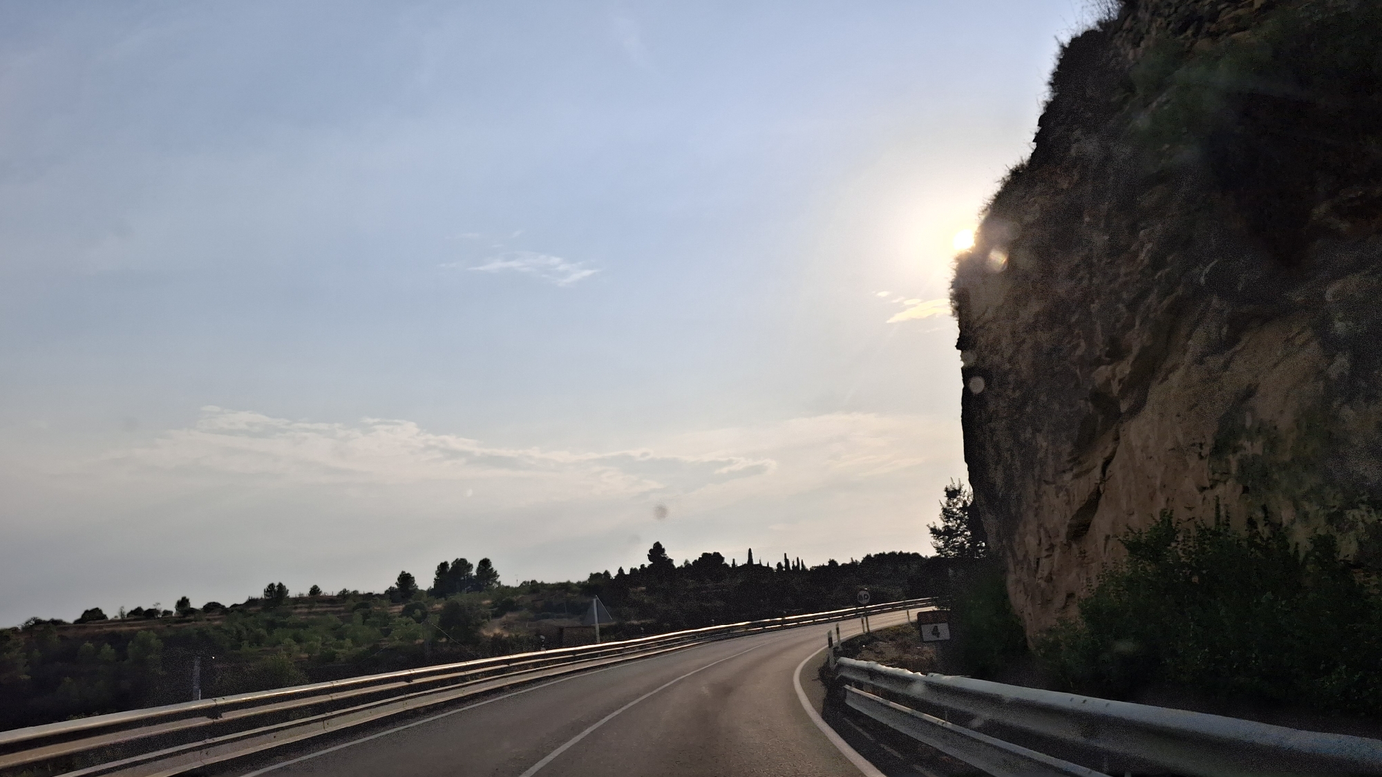A curving asphalt road passes by a steep, rocky cliff face on the right side of the frame. The road is bordered by metal guardrails on both sides. The sky is a pale blue with scattered clouds, and the sun is shining brightly, partially obscured by the top of the cliff, casting lens flares. The cliff is textured and shows signs of erosion. Vegetation, including trees and bushes, grows on the hillside to the left of the road and at the base of the cliff. A road sign with the number "4" is visible near the guardrail on the right. The scene appears to be a scenic route, likely in a rural or mountainous area.