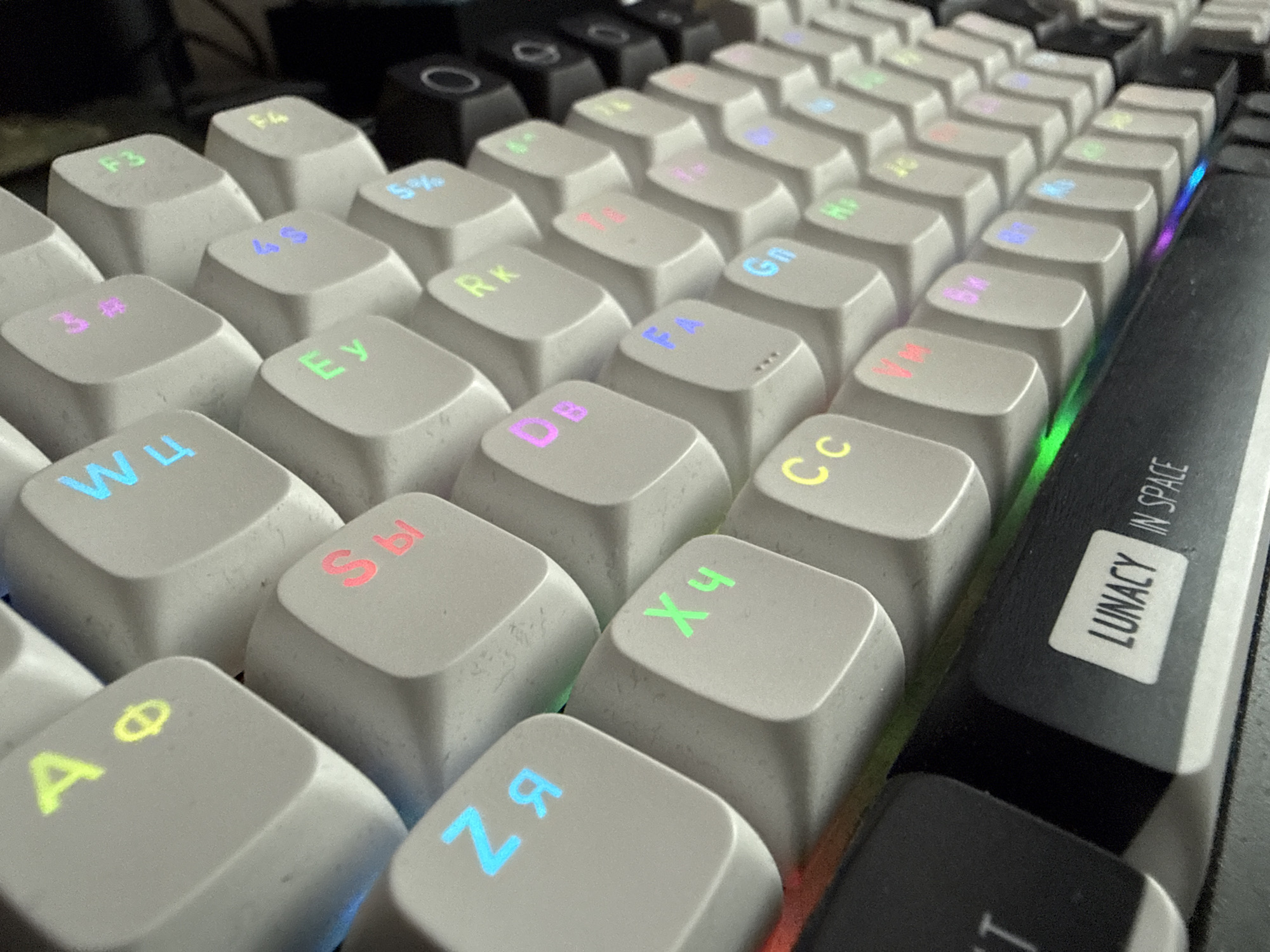 This is a close-up, high-angle, eye-level shot of a mechanical keyboard with light-colored keycaps and multi-colored backlighting. The keys are arranged in a standard QWERTY layout with some Cyrillic characters visible. The keycaps have a slightly textured surface. The lighting is artificial and casts colorful glows on the keys. In the lower right portion of the image, a black keycap with white text "LUNACY IN SPACE" is visible, suggesting this is a custom or novelty keyboard. The background is out of focus but appears to be a desk or table surface with other dark objects. There is no information in the image to indicate a location in Petropavlovsk-Kamchatsky, Russia.