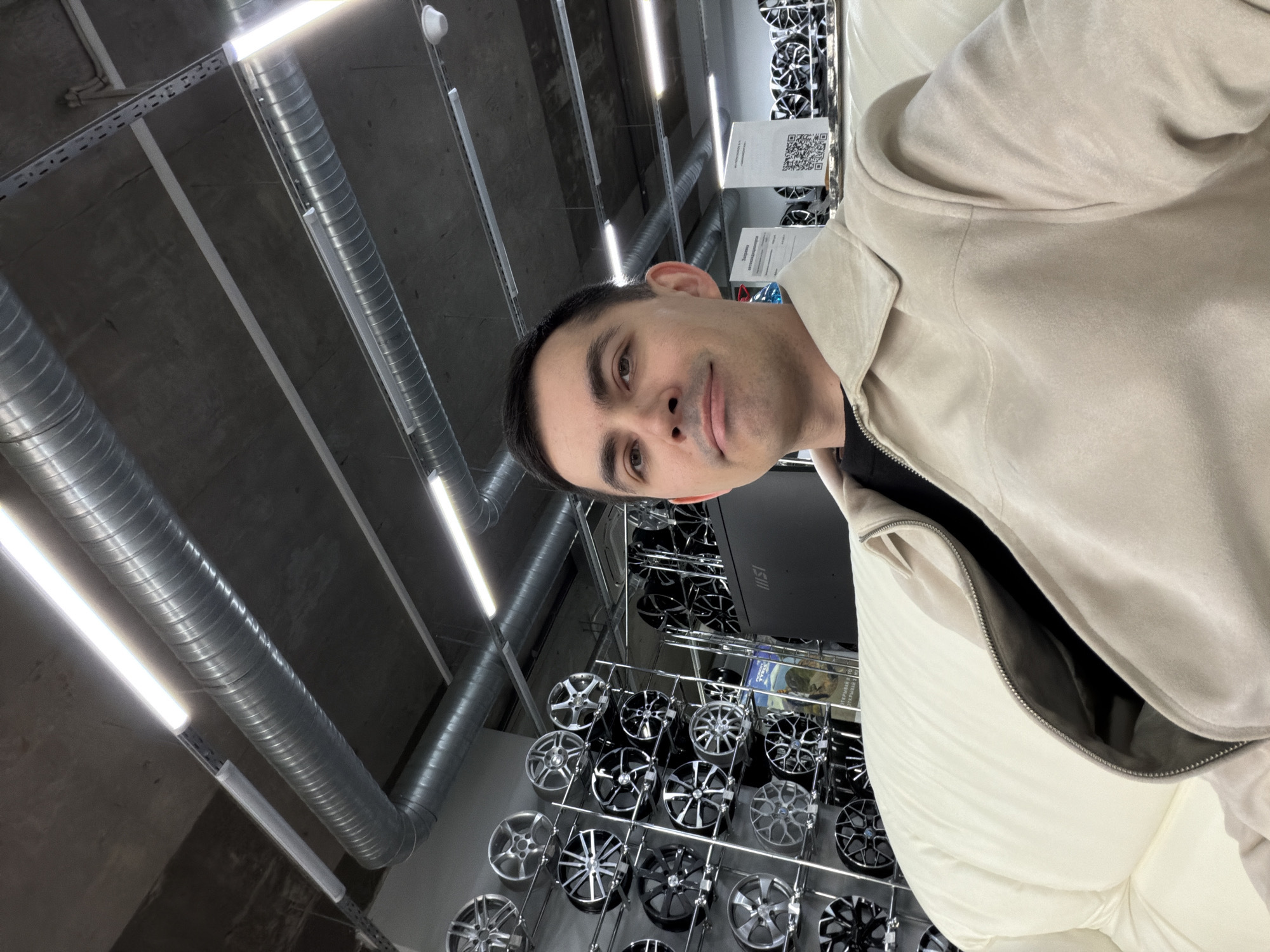 A man is standing in a store in Izhevsk, Russia, posing for a picture. He has short, dark hair and is wearing a light-colored jacket over a dark shirt. Behind him are rows of car wheels displayed on shelves. The store has exposed metal piping and bright fluorescent lighting on the ceiling. Information signs are visible amongst the wheels.