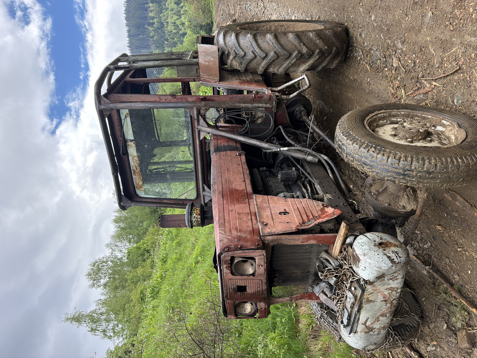 The image shows a rusty, dilapidated tractor located near Рилски манастир, Bulgaria. The tractor is missing parts and is surrounded by mud and vegetation. The environment consists of a dirt area bordering a grassy hillside with trees and a cloudy sky.