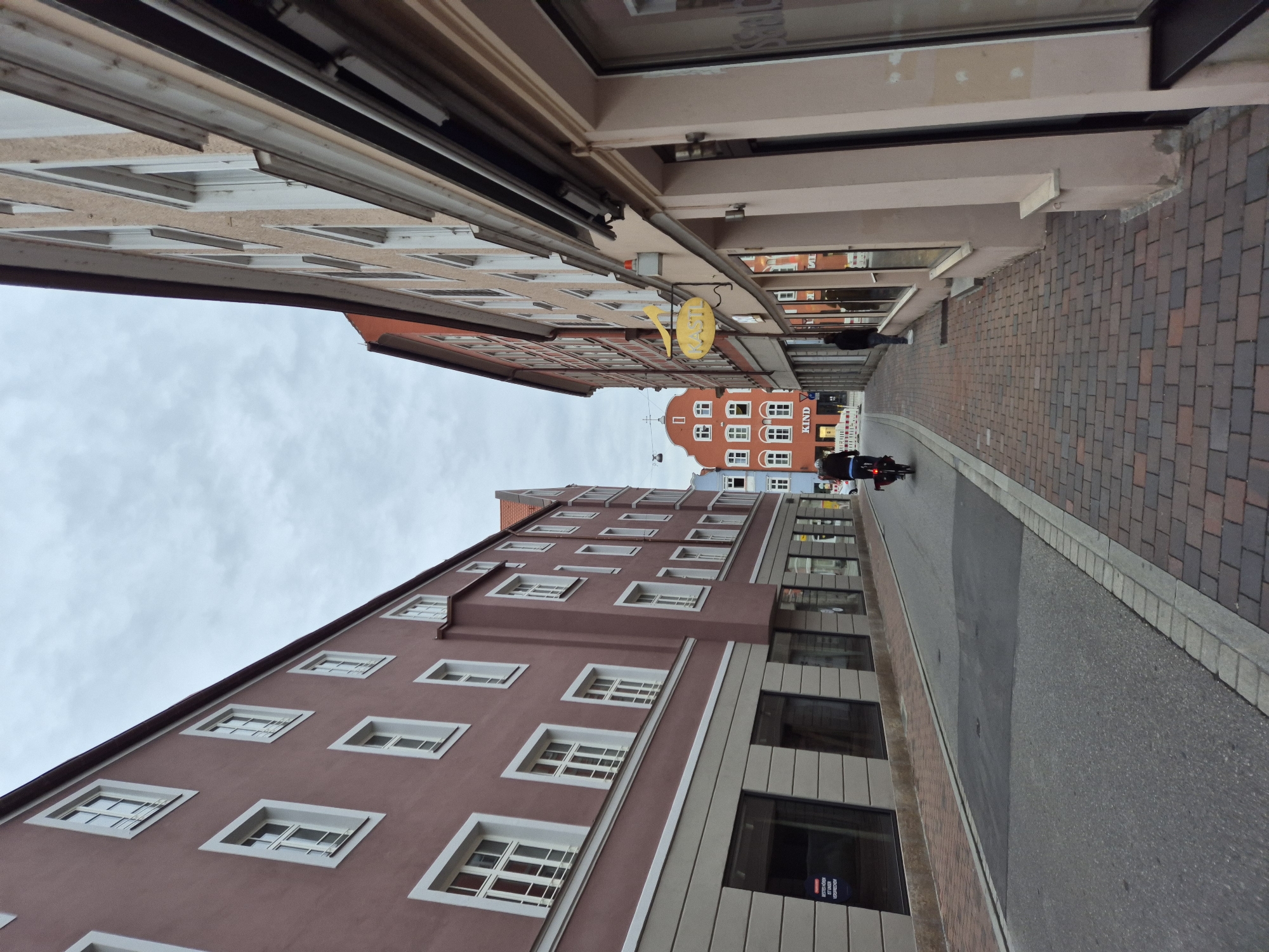 This is a street-level, slightly upward-angled medium shot of a narrow street in Landshut, Germany, likely taken on an overcast day. The street is lined with multi-story buildings on both sides. On the left, a predominantly dark pink building features numerous white-framed windows. The ground floor appears to be retail space with large, dark windows. The sidewalk on the left is paved with stone.
On the right, a pale pink building with white-framed windows also appears to have ground-floor retail spaces. A faded yellow sign with the word "RASTIL" is visible on this building. Further down the street on the right, another building with orange and white detailing can be seen.
In the middle ground, a person on a bicycle is riding away from the camera down the street. The street itself is paved with asphalt, with a brick-paved sidewalk running alongside the right-hand buildings. In the distance, a red building with a distinctive stepped gable is visible, along with a blue building. The sky is overcast, contributing to the muted lighting. There are no other significant human interactions or actions depicted.