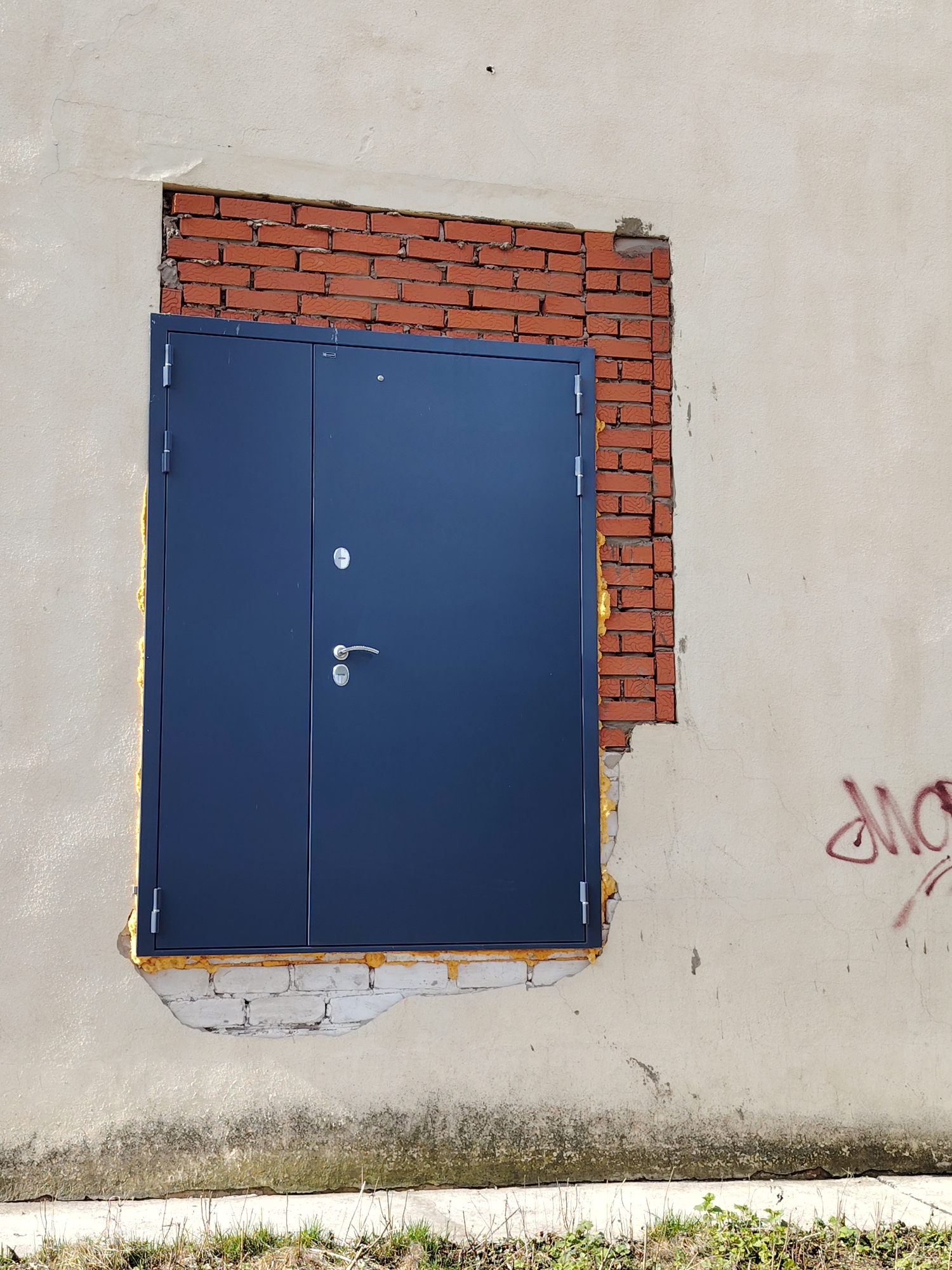 A gray metal door with a silver doorknob is set in a brick-lined opening of a light tan, textured stucco wall. The brickwork is irregularly laid with visible mortar, and the stucco around the opening is damaged and uneven. Yellow foam insulation fills the gaps between the door frame and the surrounding wall. There is faint red graffiti on the wall to the right of the door. Weeds and dry grass are visible at the base of the wall. The image was taken in the city of Chaikovsky, Russia.