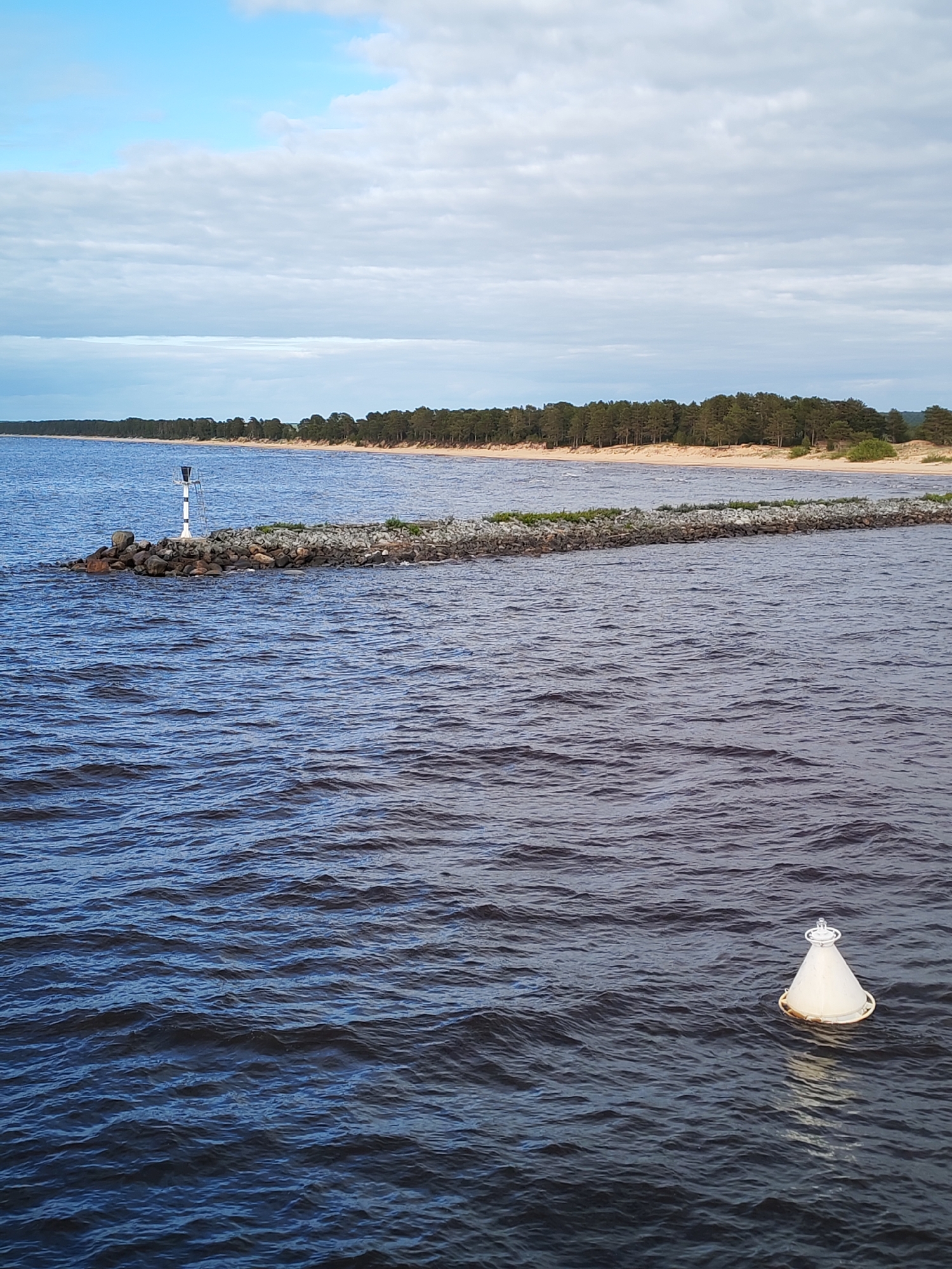The image depicts a body of water, likely a lake or sea, with a rocky breakwater extending into it. The breakwater is lined with some vegetation and a small, white and black striped navigation aid is located at its terminus. The shoreline in the background shows a sandy beach bordered by a dense line of trees. In the foreground, a white navigation buoy floats in the water. The sky is partly cloudy. The precise location in the city of Ozernoe Ust'e, Russia, is not verifiable from the visual information alone.