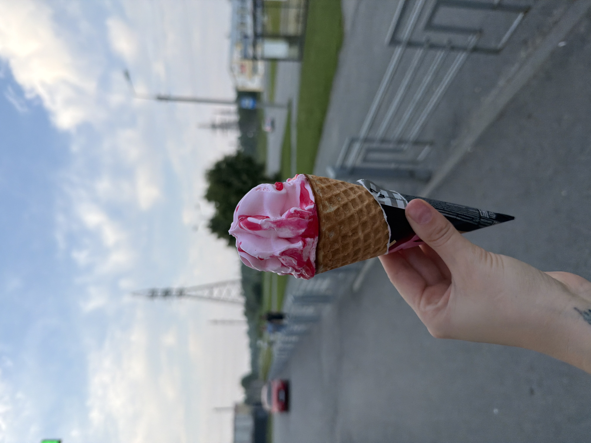 The image shows a person's hand holding a pink ice cream cone against a blurred background of a street scene. The ice cream is pink with swirls of a darker pink or red sauce. The cone is a typical waffle cone. The hand has a small tattoo on the wrist. In the background, a grey road is visible, with a red car in the distance. There are also some metal structures and trees. The sky above is cloudy and appears to be daytime. The overall impression is casual and outdoors.