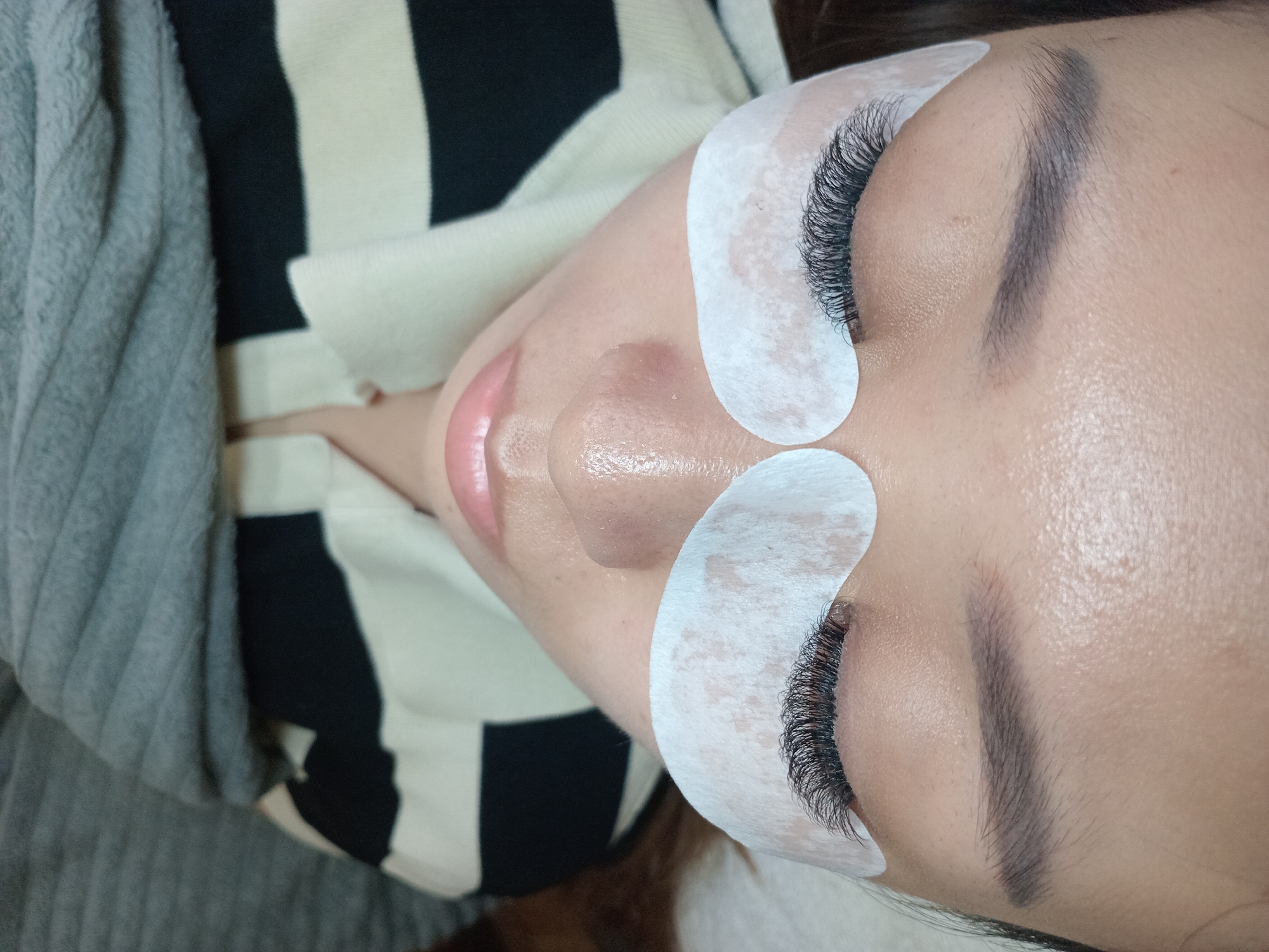 This close-up, overhead image captures a person, likely a woman, lying down with her eyes closed, suggesting she is undergoing or has just completed an eyelash extension procedure. The scene takes place indoors, with the provided context indicating North Kuta, Indonesia.
The central focus is on the person's face, primarily from the chin upwards. Both eyes feature prominent, dense, and dark black eyelash extensions, which are long and curled, indicating a professional application. White, semi-transparent under-eye pads are meticulously placed beneath each eye, typically used to protect the lower lashes and the delicate under-eye skin during the treatment.
The person's natural eyebrows are dark and well-defined. Her lips are a soft pink hue and slightly parted. Her skin appears smooth, with a subtle sheen visible around the nose area.
For attire, she is wearing a top characterized by distinct black and white horizontal stripes, visible on the upper right side of the frame. To her left, a textured grey blanket or garment is draped, contributing to a sense of comfort and a relaxed setting for the beauty treatment. There is no visible text in the image.
