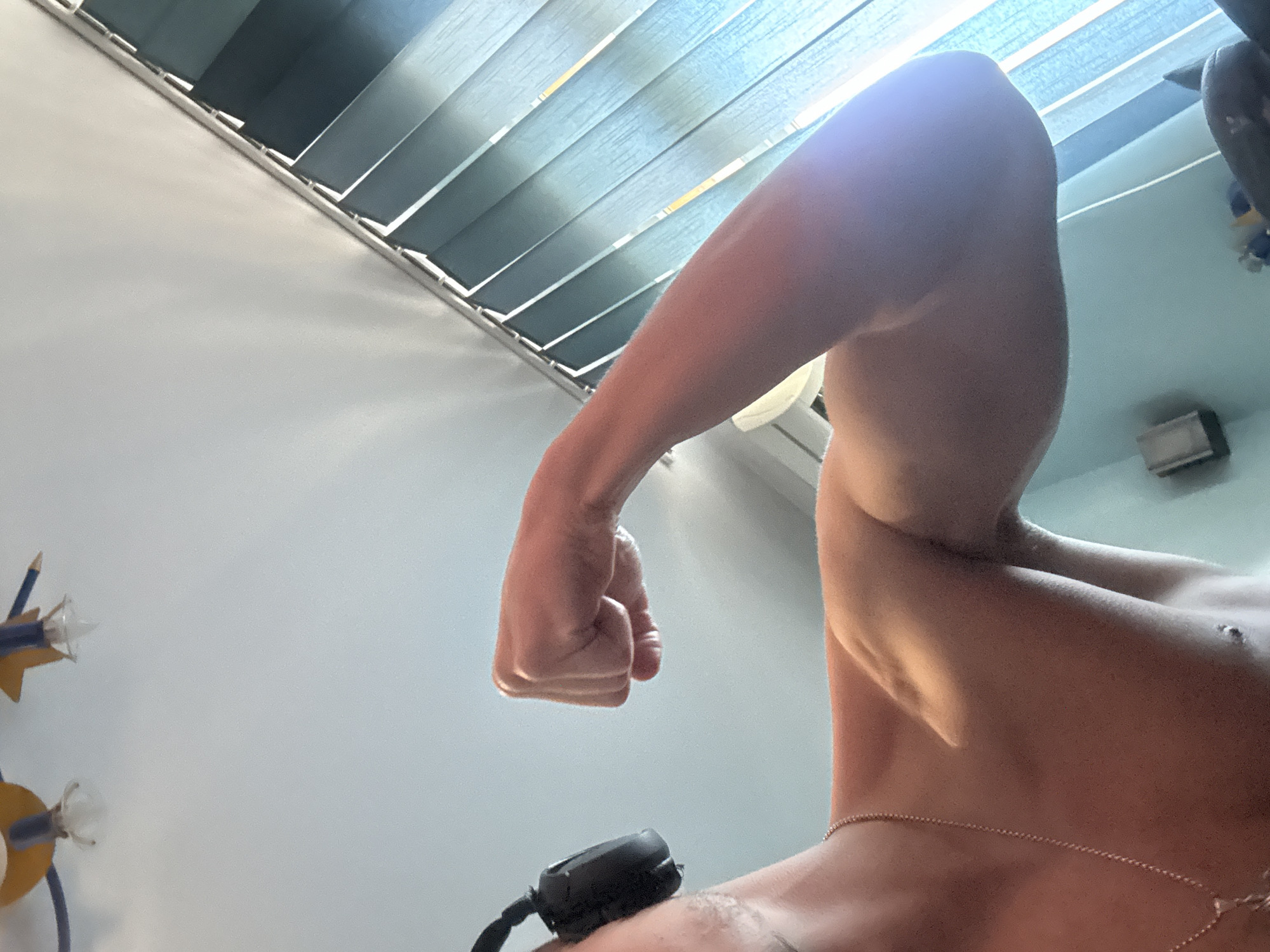 The image shows a low-angle perspective of a person flexing their right arm. The person is shirtless and a thin chain necklace is visible around their neck. A small, black device with a cord is positioned near the person's shoulder. The background includes a light-colored wall and blinds. The location is not discernible within the provided image and thus cannot be linked to Buhovo, Bulgaria.