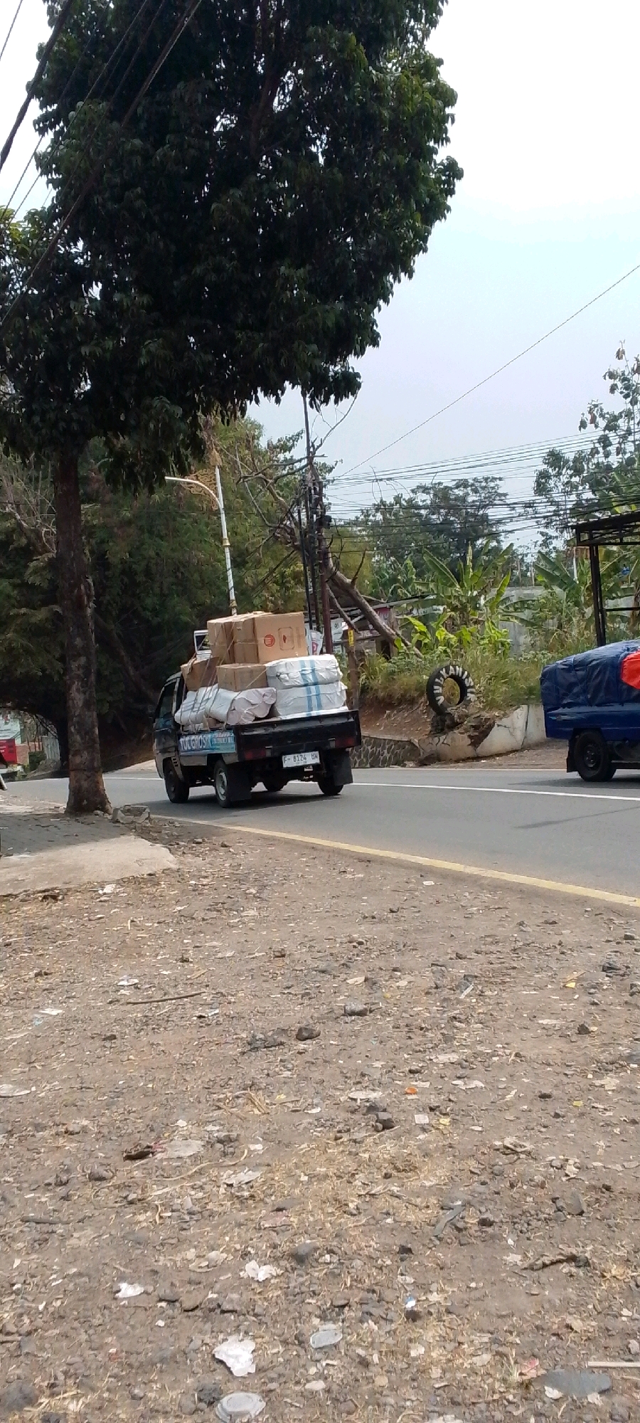 The image depicts a scene in Cilaku, Indonesia, on a road that appears to be in a semi-urban or rural area. A dark-colored utility truck, loaded with several boxes and wrapped items, is traveling away from the viewer on the right side of the road. The boxes are stacked high, with some appearing to be cardboard and others wrapped in white and blue striped material. A smaller, blue truck covered with a blue tarp is visible further down the road, also traveling in the same direction.
On the left side of the road, a large tree with dark green foliage dominates the foreground. Power lines are visible stretching across the upper portion of the image, strung between utility poles. The road surface is asphalt with a yellow dividing line. The environment includes a dirt embankment along the side of the road, littered with debris, including stones, dry grass, and various pieces of trash. In the background, there are more trees, some with banana leaves, and a few structures, including what appears to be a simple open-sided shelter or building. The sky is overcast, suggesting a cloudy or hazy day. The overall impression is of a busy but somewhat unkempt roadside environment.