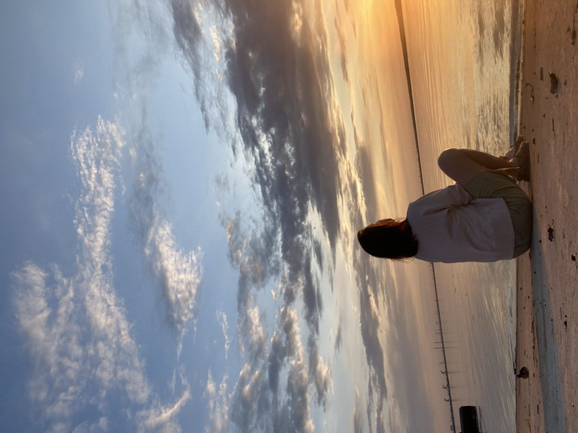 A young woman with dark hair is sitting at the edge of a body of water at dusk, likely in the city of Chaikovsky, Russia. She is wearing a light-colored shirt and dark shorts. The sky is filled with clouds. The body of water is reflective, mirroring the colors of the sky. The seating area is concrete and there are posts and pilings in the distance.