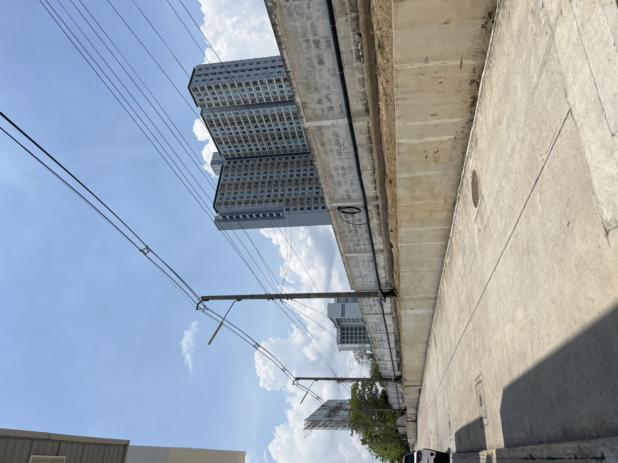 The image, taken in Pattaya City, Thailand, shows a street scene under an elevated concrete structure, likely a highway or train track. Two tall apartment buildings are visible beyond the structure, appearing gray with numerous windows. Overhead, electrical lines stretch across the sky, supported by poles. A billboard and some trees are seen further down the street. The sky is blue with scattered white clouds. The photograph is oriented with the sky to the left and the street to the right.