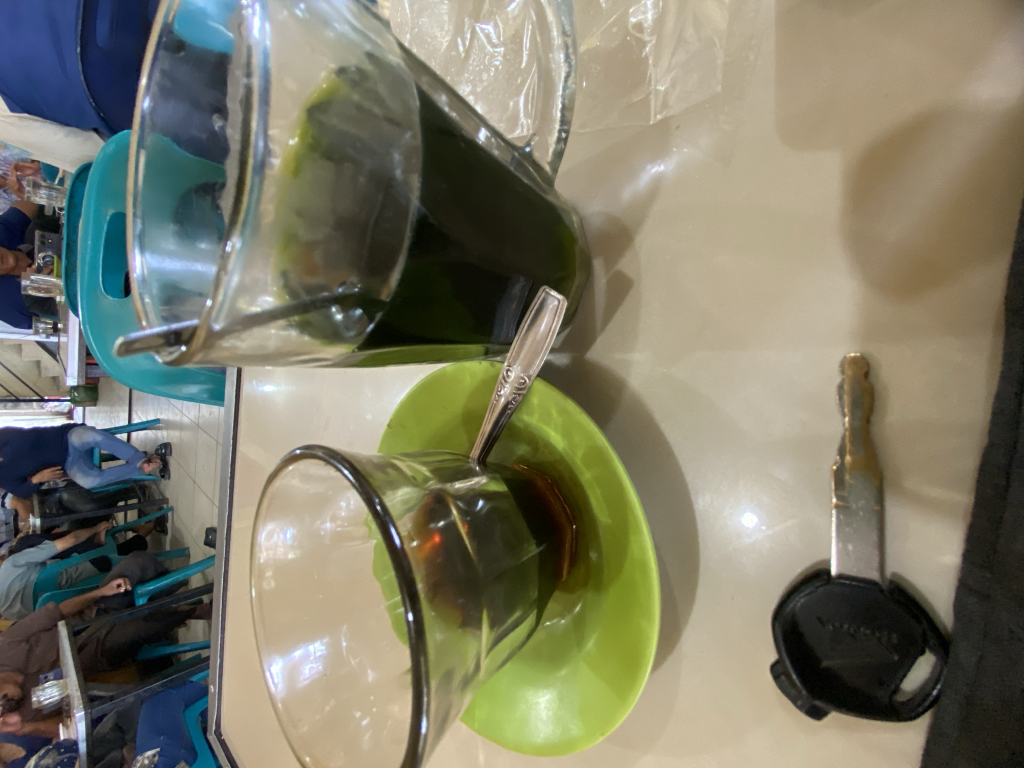The image depicts a casual setting, possibly a warung kopi or a local eatery, in Lhokseumawe, Indonesia. In the foreground, a car key with a black head is lying on a table. To the right of the key, a glass of dark green liquid, likely a beverage, is placed next to a clear glass containing a dark brown liquid, possibly coffee. A small green saucer with a spoon is underneath the clear glass. In the background, several individuals are visible, seated at tables. One person is wearing a brown long-sleeved shirt and dark pants, another a light blue shirt and jeans, and a third a dark blue polo shirt and jeans. The environment suggests a relaxed, communal space, common in Indonesian cities.