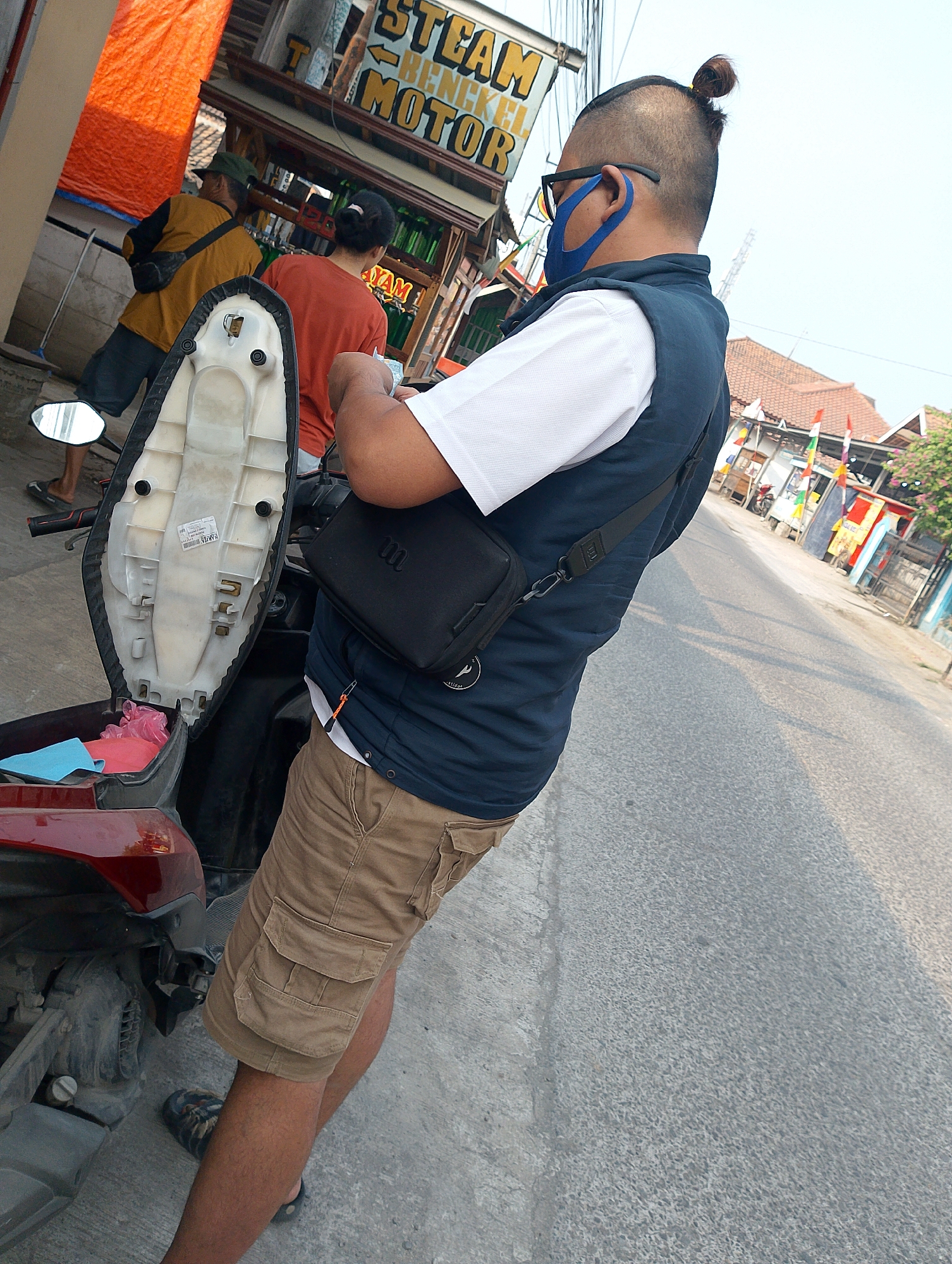 A man with a bun hairstyle and a blue face mask is standing next to a motorcycle in a street scene in Karawang, Indonesia. He is wearing a white shirt, a dark blue vest, and khaki cargo shorts. A black sling bag is worn across his chest. The motorcycle's seat is open, revealing storage underneath. In the background, other individuals are visible near a building with signage that appears to be for a business. The street is paved, and flags are displayed along the buildings. The overall impression is a casual street-side interaction or stop.