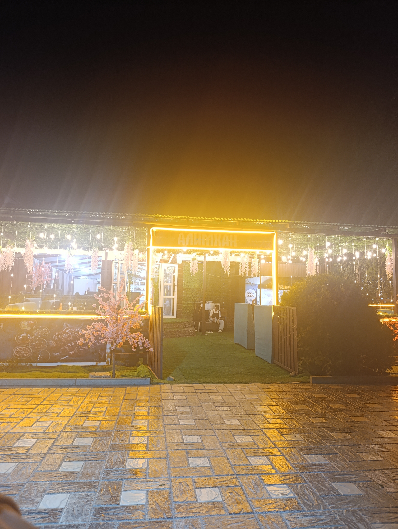 The image depicts an outdoor seating area of a restaurant or cafe in Bosteri, Kyrgyzstan, at night. The establishment features a covered patio adorned with string lights and decorative hanging flowers. A neon sign with Cyrillic lettering, possibly the name of the establishment, is prominently displayed in orange. Artificial cherry blossom trees add to the ambiance. Inside the covered area, it appears to be a dining space with tables and chairs, and possibly a bar visible. In the foreground, a person is seated on a box. The ground is paved with patterned tiles, which are wet and reflect the surrounding lights, suggesting recent rain. To the right, there is a fenced-off area with lush green grass and a dense bush. The overall impression is of a well-lit and inviting outdoor venue.