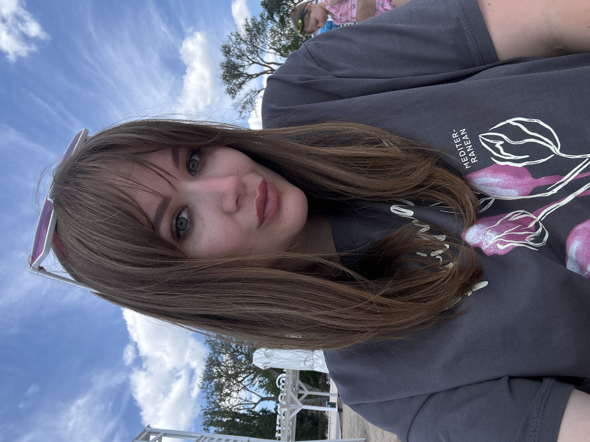 The image presents a woman with fair skin, light brown hair, and blue eyes, posing in front of a clear blue sky with scattered white clouds. She wears a dark gray t-shirt with a graphic design that includes white and pink floral elements, along with the word "MEDITER-RANEAN". The lower portion of the image reveals white structures, possibly gazebos or architectural elements, suggesting an outdoor setting near the beach. A tree is partially visible behind the structures. The location is likely Yevpatoriya, Ukraine, based on visual cues and the prompt's request. In the background, a child is wearing a pink bathing suit.