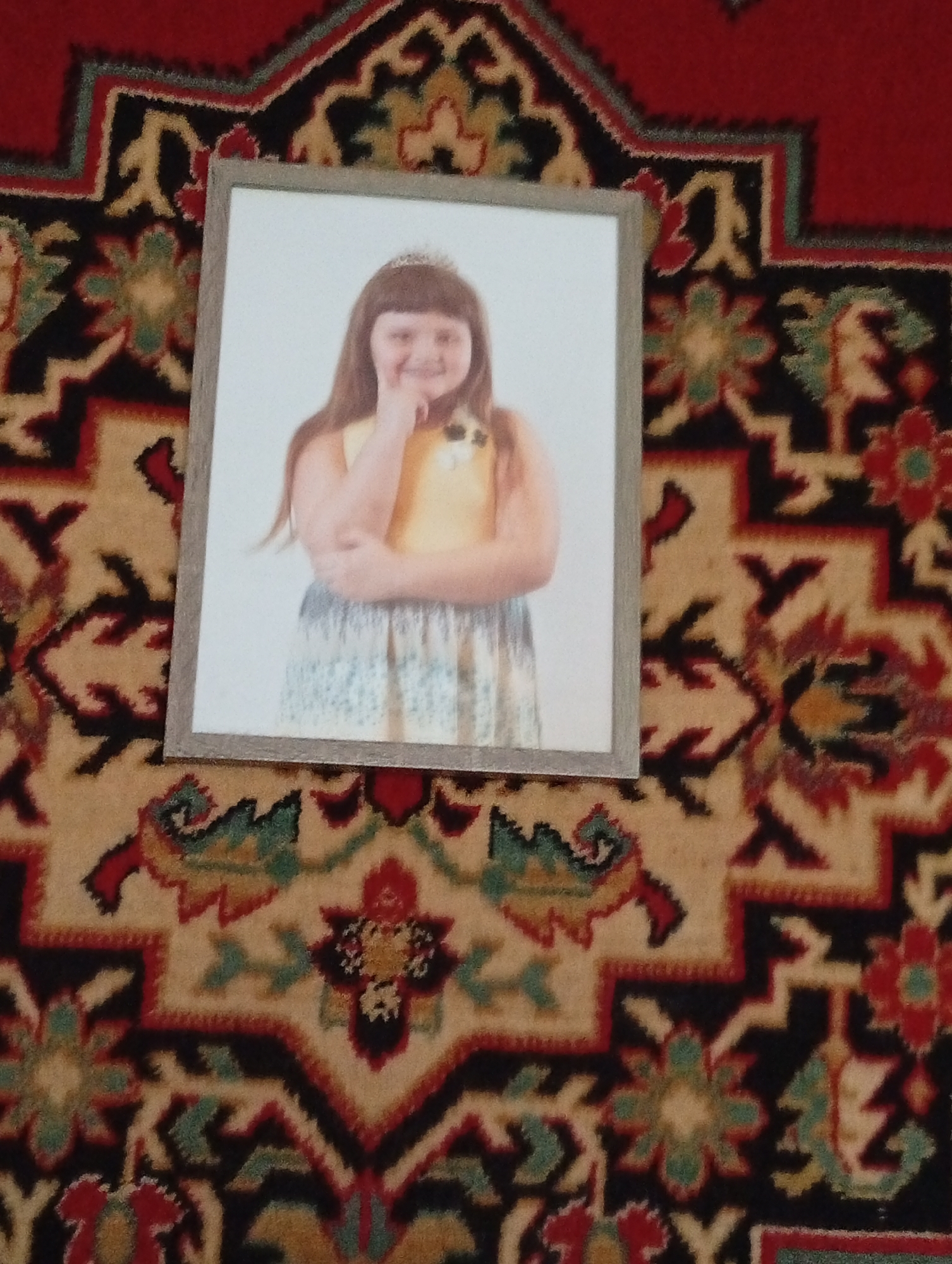 A framed photograph of a young girl wearing a tiara and a yellow and blue dress lies on a red, beige, and black patterned rug. The girl is smiling and has her hand on her chin. The rug appears to be a traditional Pavlovsky Posad shawl or carpet, characteristic of the city of Pavlovsky Posad, Russia, known for its textile production. The image is taken from a high angle, with the rug filling most of the frame.