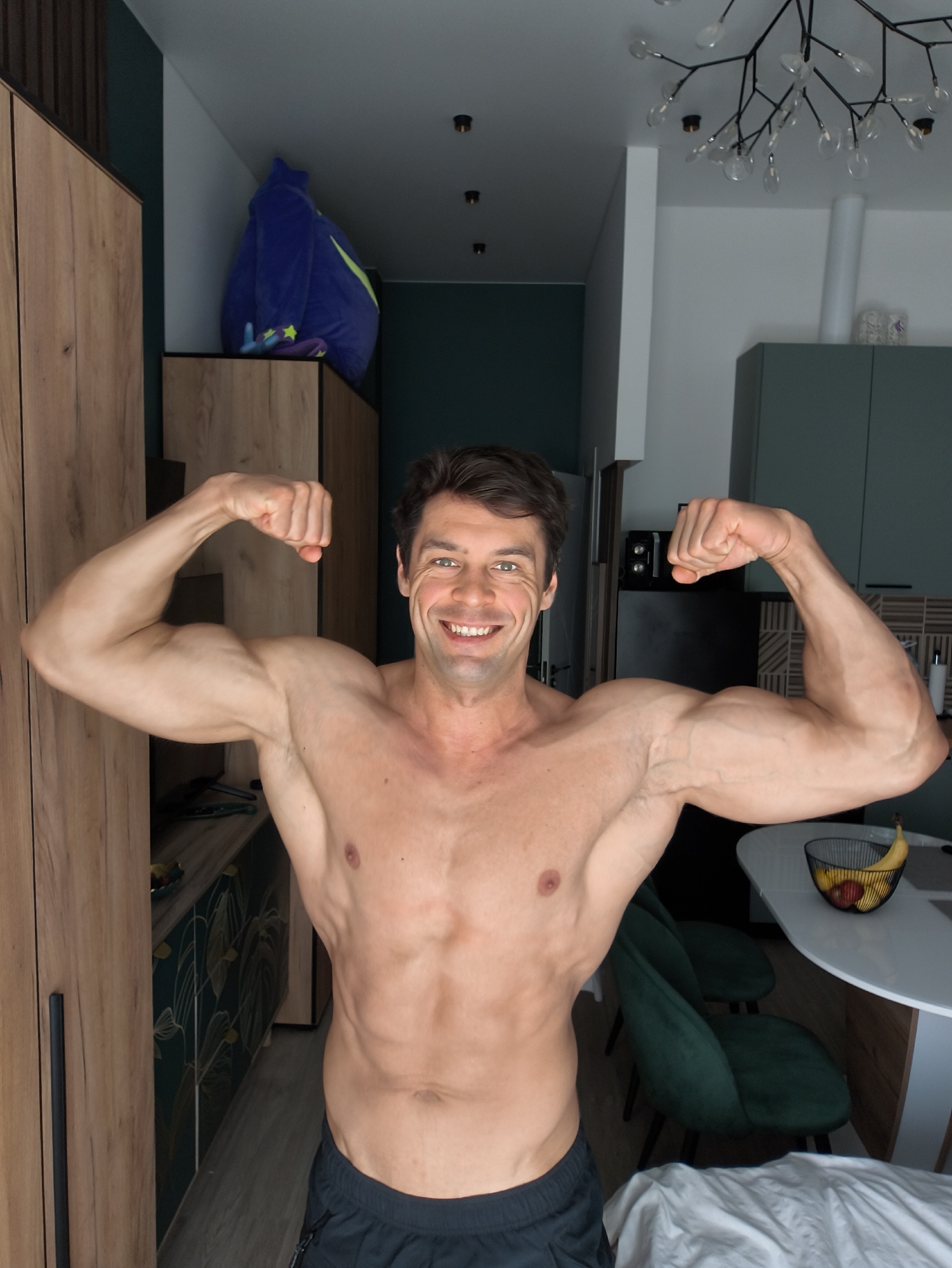 A shirtless man, presumably in his thirties, stands centered in the frame, flexing both arms to display his bicep muscles. He smiles at the camera. The setting appears to be the interior of an apartment, possibly in Moscow, Russia, based on the modern design and general aesthetic. Background elements include kitchen cabinets with decorative leaf patterns and a round table with a fruit bowl containing bananas and apples. A large, possibly bean bag chair is situated atop a closet or cabinet unit in the background. Overhead, a modern chandelier is visible. The lighting is natural.