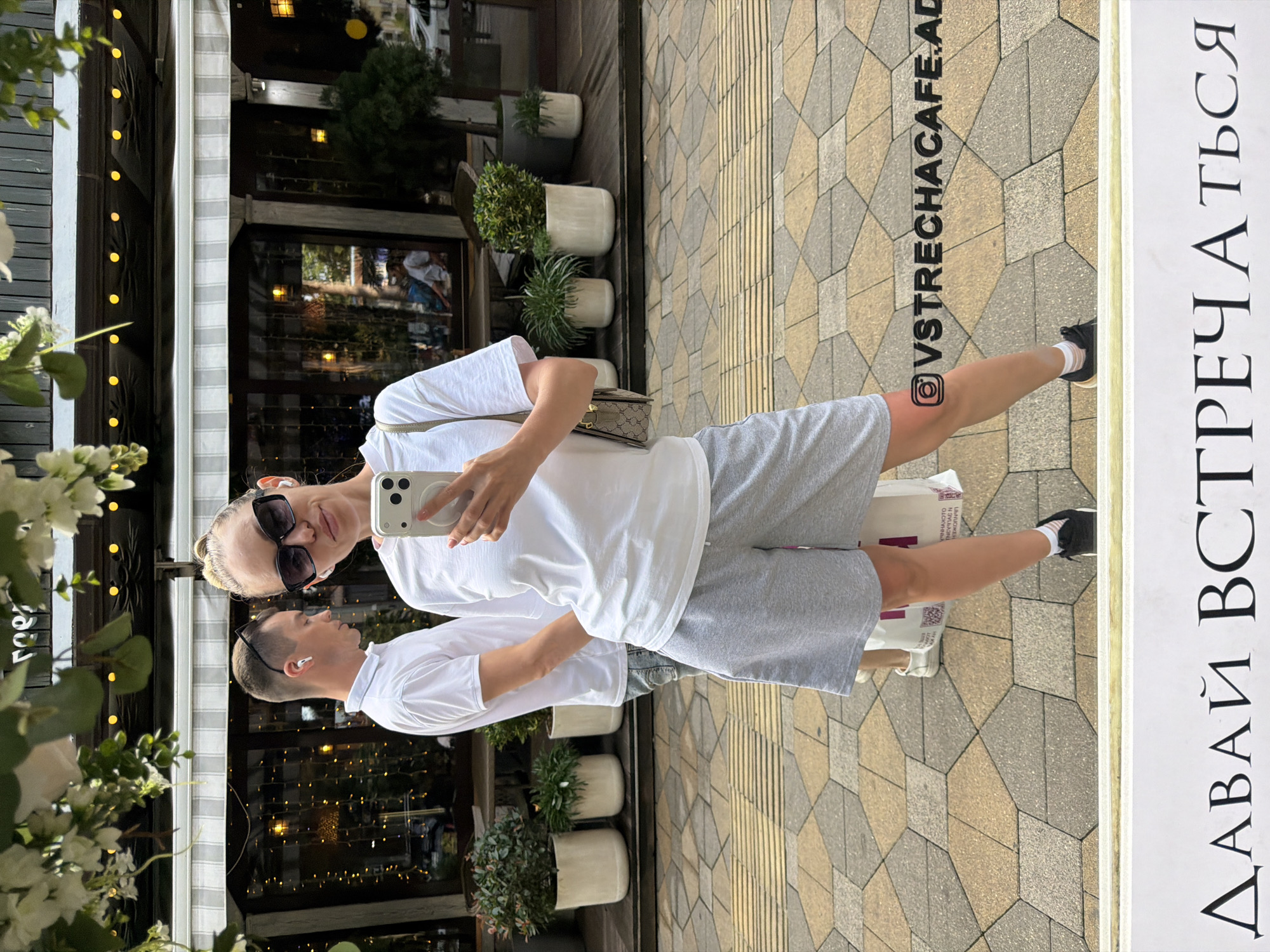The image depicts a woman taking a selfie in front of a cafe in Sochi, Russia. She is wearing a white t-shirt, grey shorts, sunglasses, and black sneakers. A man in a white polo shirt stands behind her. The cafe appears to be on a street with a cobblestone sidewalk. The weather is overcast, and there are plants and flowers visible in the foreground. A social media handle, @VSTRECHACAFE.AD, is overlaid on the image. The Russian phrase "ДАВАЙ ВСТРЕЧАТЬСЯ" (Let's meet) is visible at the bottom of the image.