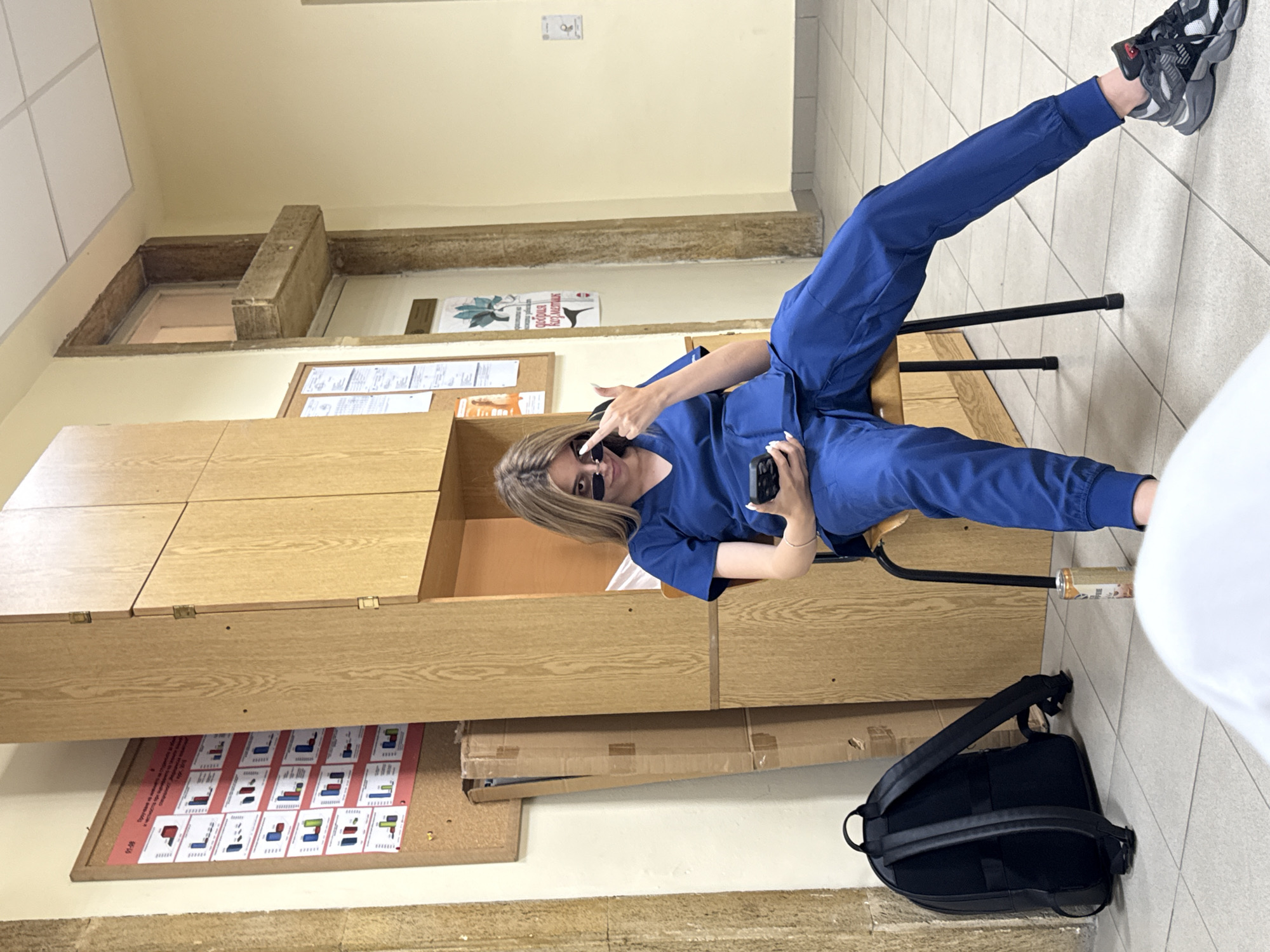 A young woman in blue scrubs sits in a chair in a well-lit room, with one leg extended towards the upper right corner of the frame. She wears sunglasses and holds a phone in her left hand, while gesturing with her right hand. A backpack sits on the floor to her right. The room has tiled floors, beige walls, and a built-in wooden cabinet system. There are posters and informational sheets attached to the walls and cabinets. Based on the style of the building and informational sheets, the location appears to be in the city of Pleven, Bulgaria, possibly within a medical or educational facility.