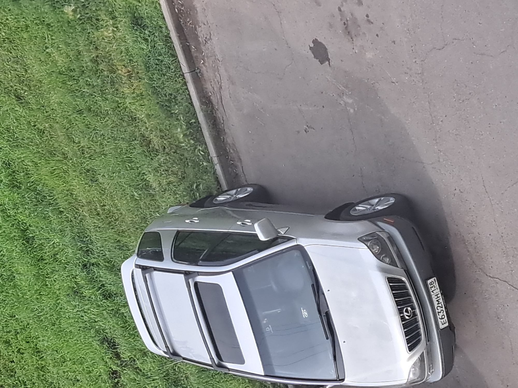 The image shows a silver Lexus RX 300 SUV parked on a paved surface adjacent to a grassy area with a concrete curb. The car's license plate is visible and reads "E632MH 138 RUS," indicating it is registered in the Irkutsk region of Russia. The car has a sunroof and roof rails. The scene appears to be an outdoor parking area in an urban or suburban environment. No people are visible in the image.