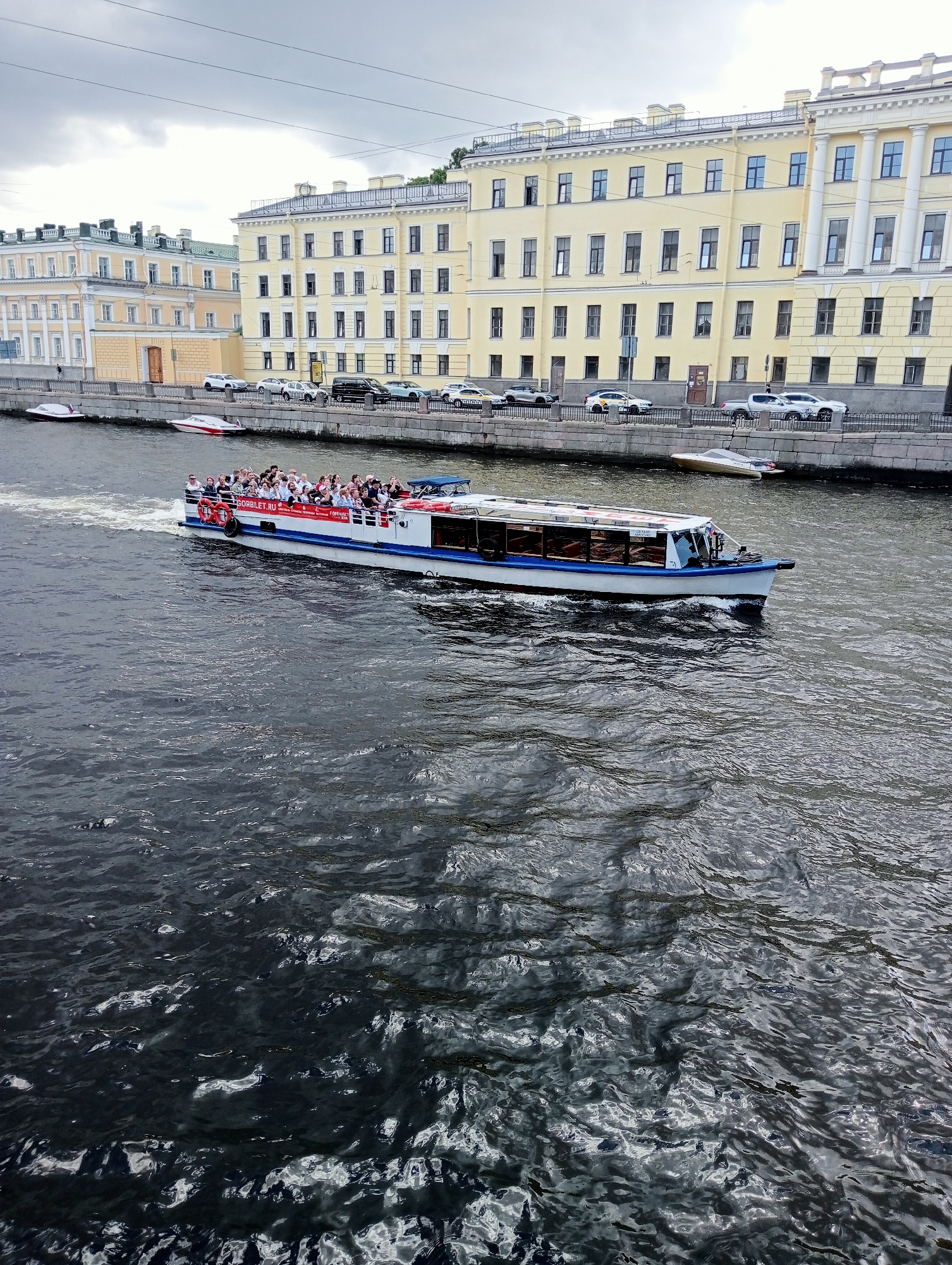 This is a medium-angle, eye-level shot of a canal in Saint Petersburg, Russia. A large white passenger boat with a blue stripe is traveling away from the camera, leaving a wake. The boat is filled with passengers standing on the upper deck and seated inside. The boat has the text "BORBILET.RU" and "TOPREMO" printed on its side. On the left bank of the canal, there are several buildings with yellow facades and white trim, characteristic of St. Petersburg architecture. Cars are parked along the embankment, and there are also two smaller boats docked along the wall. The canal water is dark and choppy. The sky is overcast with clouds.