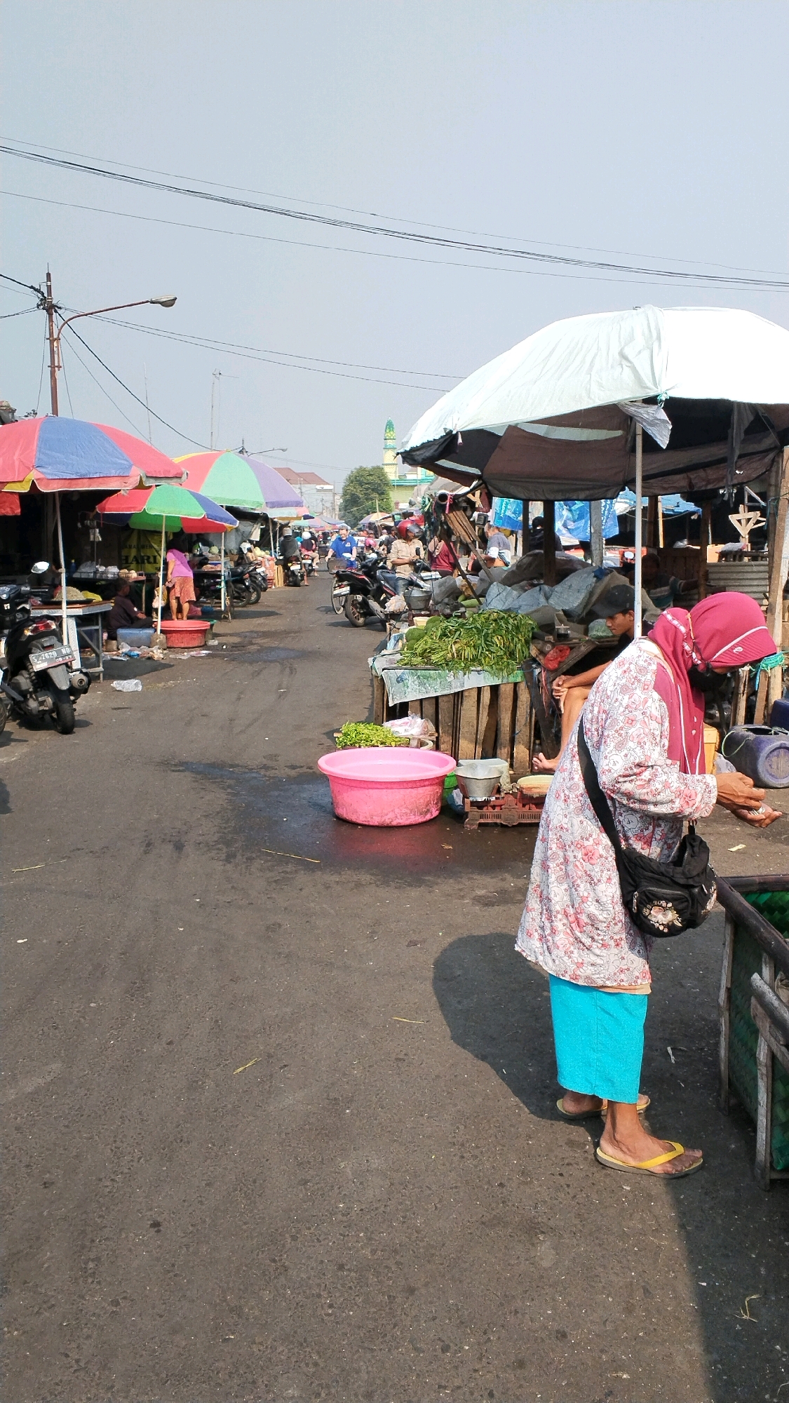 A busy outdoor market in the city of Mojokerto, Indonesia. The market is lined with various stalls, many covered by colorful umbrellas. People are seen browsing and purchasing goods. In the foreground, a woman in a pink headscarf and a patterned long-sleeved shirt stands near a stall selling fresh produce, holding some small items in her hands. She is wearing a blue skirt and yellow flip-flops. Several motorcycles are parked along the side of the road. In the background, more stalls and a mosque's minaret are visible. The ground is paved with asphalt, and there are some puddles of water. The sky is clear and bright.