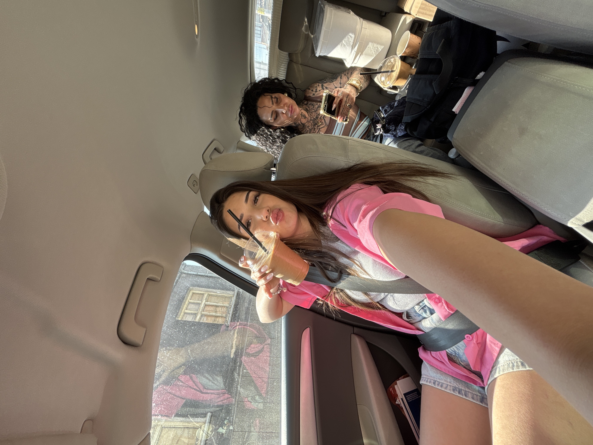 The scene is set inside a car in Rakovski, Bulgaria, illuminated by sunlight. In the front passenger seat, a young woman with long dark hair, wearing a pink open shirt over a light grey ribbed tank top and denim shorts, holds a clear plastic cup with a light brown beverage and a black straw. She is looking towards the camera with a pouting expression. A black seatbelt crosses her chest and lap.
In the rear passenger seat, a second woman with dark, curly hair and extensive tattoos visible on her arms, chest, and neck is engrossed in a gold-colored smartphone, holding it with both hands. She wears a striped top.
The car's interior features light beige headliner and seats, along with grey and lighter-toned door panels. Through the front passenger window, old buildings with faded, possibly peeling paint and multiple windows are visible, one featuring a large red and black mural or graffiti. The car window appears to have some specks or dust. On the floor behind the rear passenger seat, two white plastic storage containers are stacked, alongside a black bag and additional beverage cups (one clear plastic, one paper). A water bottle is also visible on the floor.