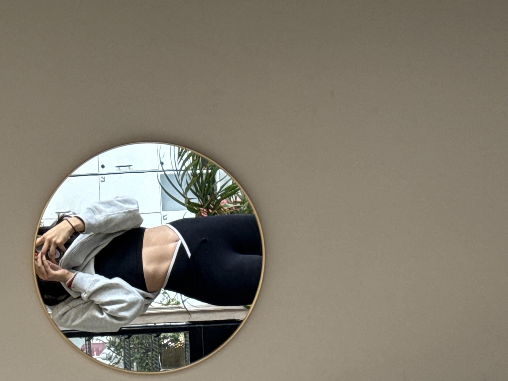 The image displays an unusual, rotated orientation, with a large, plain, light brown wall occupying most of the frame. In the lower-left section, a circular mirror with a gold-toned frame is visible, reflecting a scene that also appears rotated approximately 90 degrees counter-clockwise.
Inside the mirror, a person is shown from the chest to the upper thighs. They are wearing a black sports bra or crop top, black high-waisted leggings with distinctive thin white stripes or straps around the waist and hips, and a light grey hooded sweatshirt (hoodie) worn open. Their midsection reveals visible muscle definition. The person's face is obscured by their hands, which are holding a smartphone, indicating they are in the process of taking a mirror selfie. Dark hair is visible around their hands.
The background reflected in the mirror suggests an indoor setting in Puebla, Mexico. On the left side of the reflection, a light-colored wall features horizontal lines and faint numbers, specifically '2' and '3', which might be part of lockers or shelving units. To the right of the person, a large, leafy green plant, resembling an indoor palm, is visible. Below the person's reflection, a darker area hints at another part of the room or an outdoor view through a window, showing greenery and a structural element like a railing. The lighting suggests it is daytime. No discernible emotions are conveyed as the person's face is hidden.