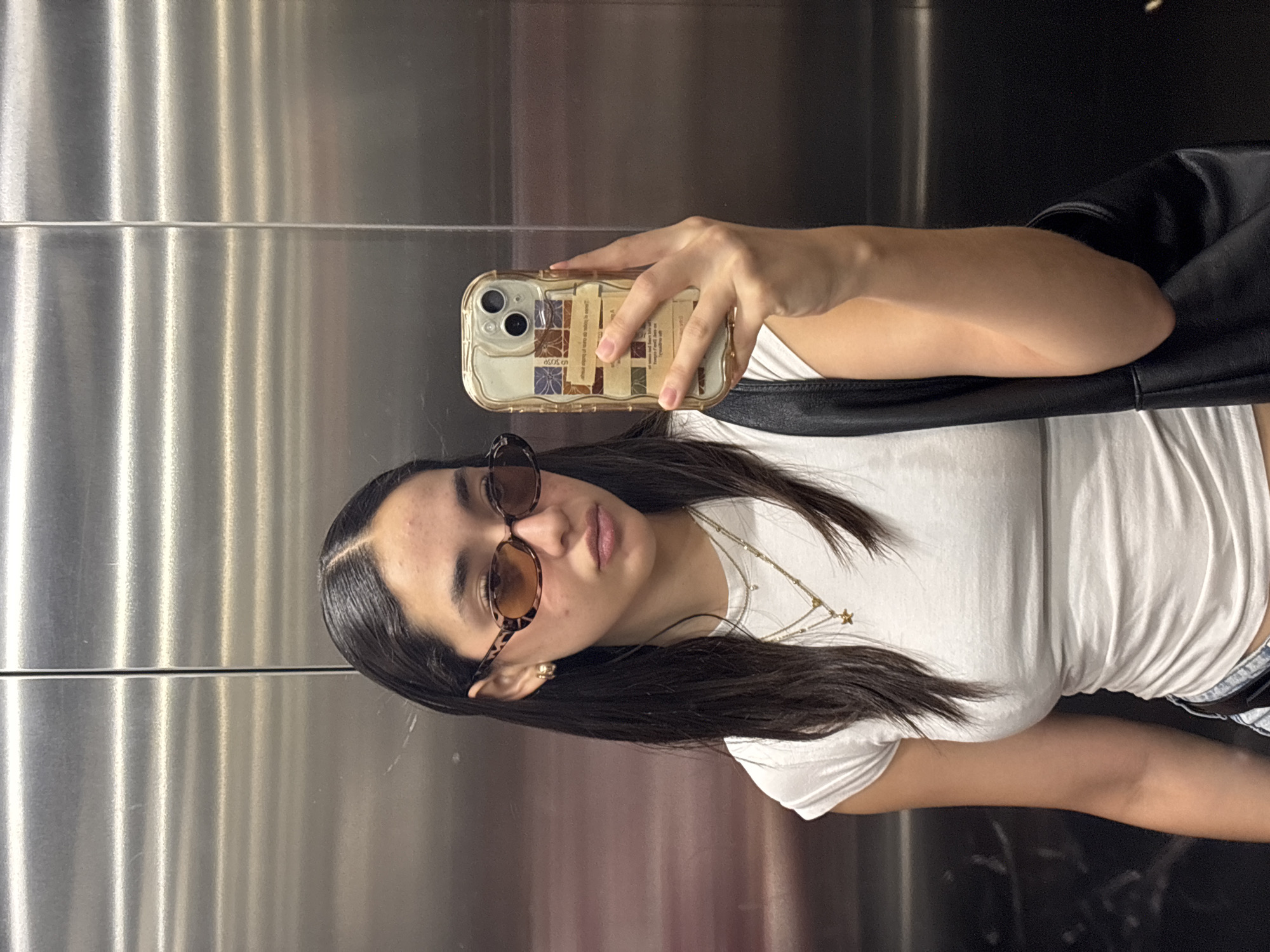 A woman is taking a selfie in what appears to be an elevator. The elevator has stainless steel walls. The woman is wearing sunglasses, a white t-shirt, a necklace, and possibly denim shorts. She is holding a phone with a decorative case. A black bag is slung over her shoulder. The location cannot be determined based on the image provided. San Andrés Cholula, Mexico is not ascertainable from the image.