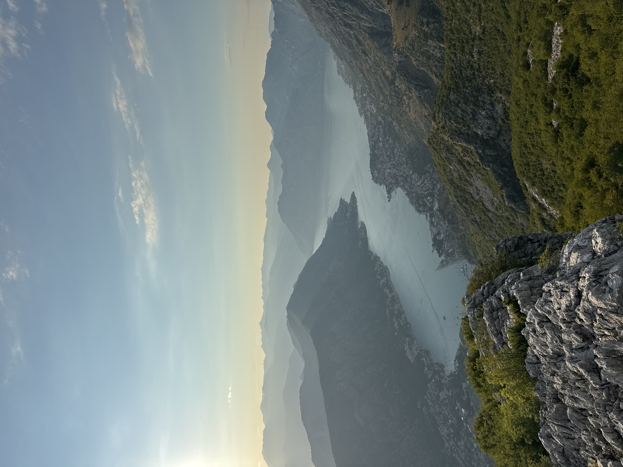 This image captures a scenic view of the Bay of Kotor from a mountainous vantage point, likely in the vicinity of Žanjev Do, Montenegro. The foreground features a rugged, grey rock formation covered with sparse green vegetation. Beyond this, the terrain slopes down, densely covered with lush green trees. In the mid-ground, the expansive, calm waters of the Bay of Kotor are visible, with a town or village nestled along its coastline. The water reflects the soft, hazy light of the sky, which transitions from a pale yellow near the horizon to a clear blue above, with wispy white clouds scattered across the upper portion of the frame. A bright sun is visible on the far left, casting a gentle glare and atmospheric haze over the distant, layered mountain ranges that frame the bay. The overall scene conveys a sense of natural beauty and tranquility, with no people or specific actions depicted.