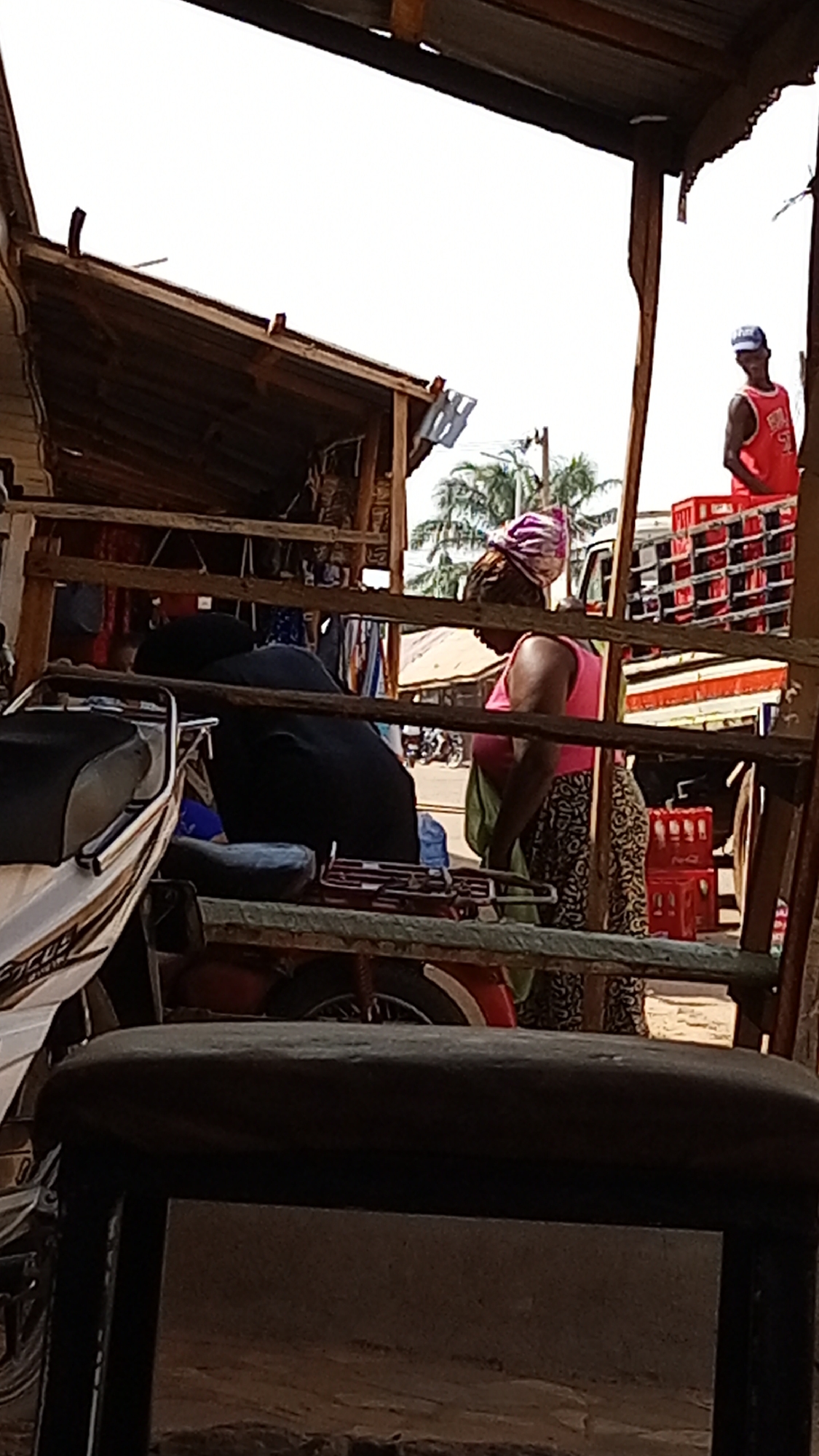 The image shows a street scene in an urban environment, possibly a market area in Unknown, Unknown. A woman wearing a pink top, patterned skirt, and a headwrap is standing near a parked motorcycle and an old wooden structure. A man in a red jersey and blue cap is visible standing on a truck loaded with red crates in the background. The scene is viewed through what appears to be part of a wooden frame or structure, possibly a stall or shop. The lighting indicates it is daytime. The focus is somewhat soft, and the foreground is partially obscured by a dark object, possibly a chair or part of the structure.