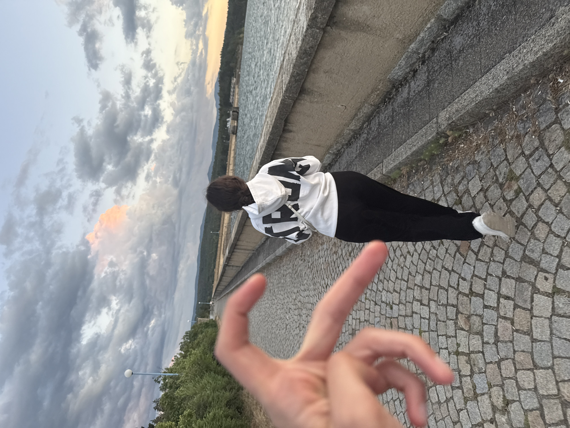 A person is walking away from the camera on a cobblestone path that runs alongside a large body of water. The person is wearing a white hooded sweatshirt with black lettering and black pants. The body of water is bordered by a stone wall. In the background, there are trees and hills, with a dam structure visible in the distance. The sky is filled with dramatic clouds, with hints of pink and orange from a sunset. In the foreground, a hand is held up, forming a heart shape. This scene is located at the Koprinka Dam in Koprinka, Bulgaria.