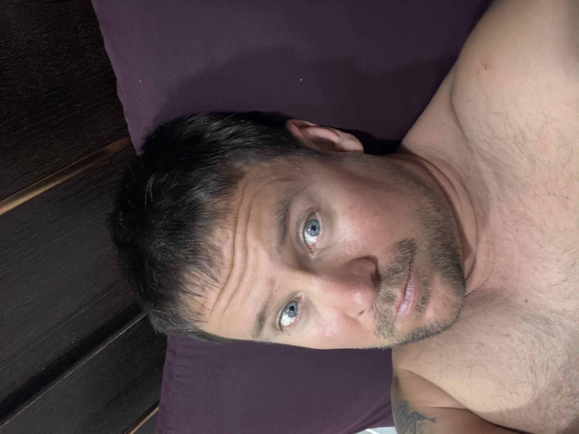 A shirtless man with blue eyes and light brown hair is lying down. He has stubble on his face and a small tattoo on his left shoulder. The background appears to be a bed with a dark wooden headboard and a purple pillow. The lighting is dim. The image does not provide any information about the location in the city of Baan Mae Nam, Thailand.