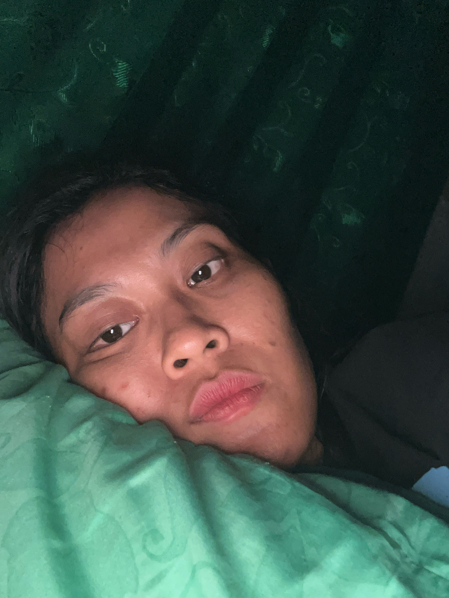 The image is a close-up, low-angle selfie of a person lying down. The person has dark hair and brown eyes, and their head is resting on a green pillow. The background appears to be a dark green curtain. The overall lighting is dim. The image is not geo-tagged, so it cannot be located in Kecamatan Pati, Indonesia.