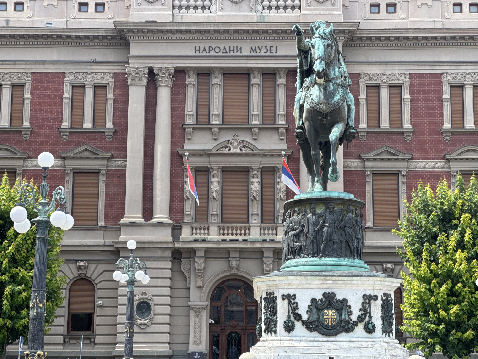 The image shows a wide shot of the National Museum in Belgrade, Serbia. In the foreground is a large bronze equestrian statue of Prince Mihailo. The statue is on a tall, ornate stone pedestal with bas-reliefs depicting historical figures and Serbian coats of arms. Two Serbian flags are visible on poles in front of the museum. The museum building itself is made of red brick with stone accents and classical architectural features, including columns and arched windows. There are trees on either side of the statue and lamp posts on the left and right. The sky is not visible.