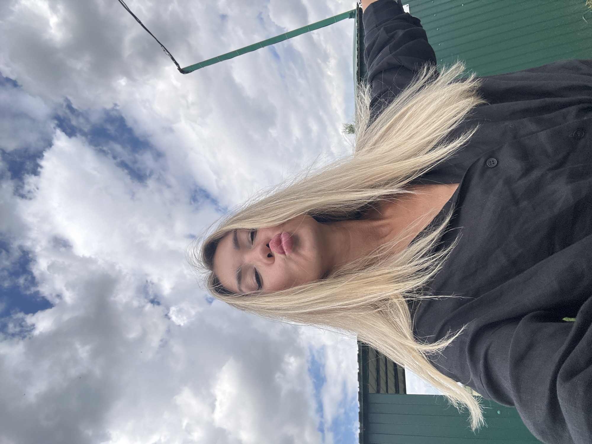 A young woman with blonde hair is captured in a selfie outdoors. She is wearing a black button-up shirt and puckering her lips. The background features a cloudy sky, a green building, and a tall green pole with a wire attached. The overall setting appears to be a rural or semi-urban area, possibly on the outskirts of Фельдмаршальский, Russia.