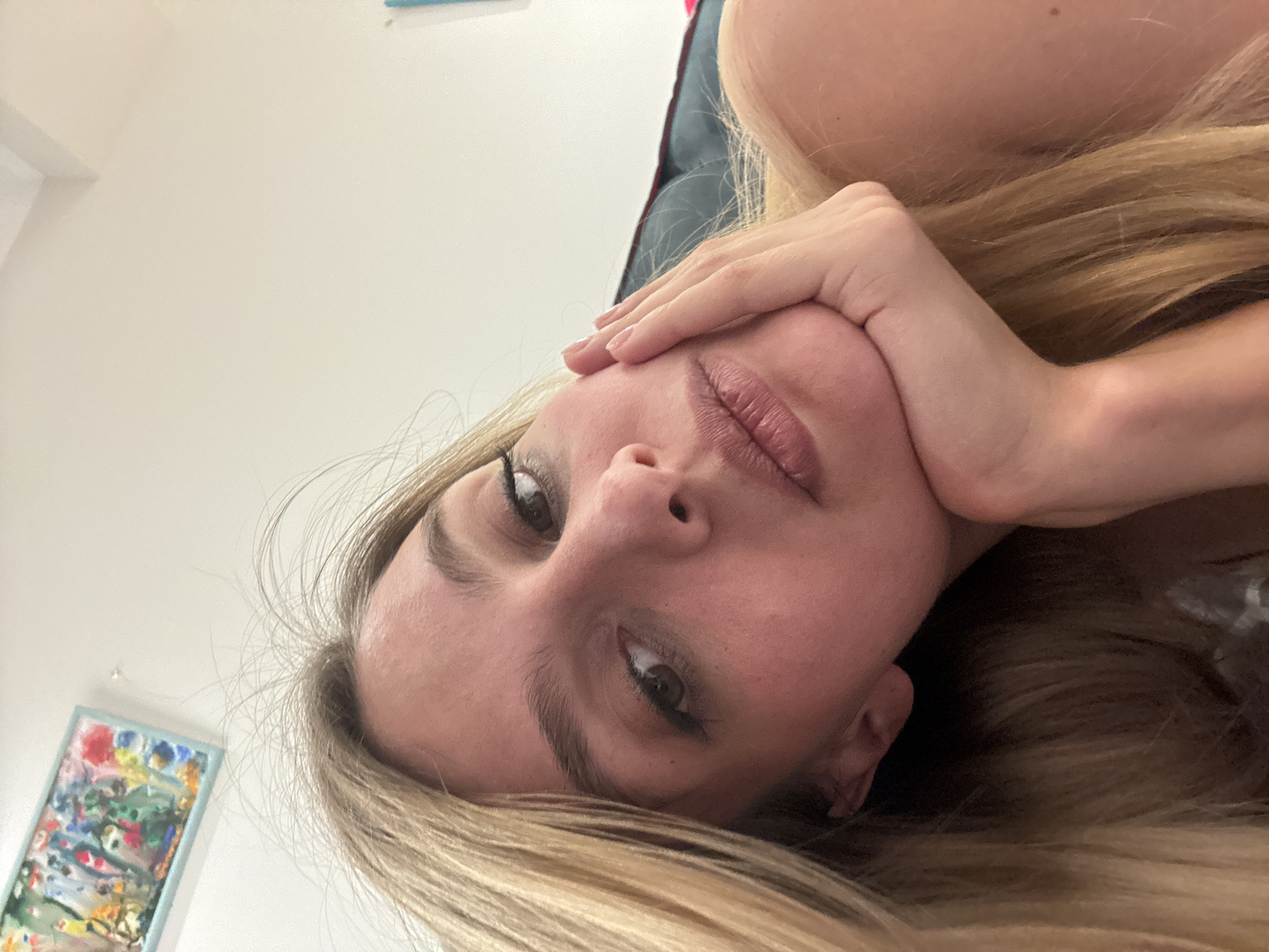 The image features a close-up, rotated portrait of a person, likely a woman, with long blonde hair. She is looking directly at the camera from what appears to be a low angle, suggesting she is lying down. Her right hand (viewer's left) rests gently on her cheek and forehead, with manicured nails visible. Her lips are slightly parted, and her eyes are open, with subtle eyeliner enhancing her gaze.
The background is predominantly a plain, light-colored wall, likely white or off-white. In the lower-left corner, a framed abstract painting with a light blue or teal border is visible. The artwork contains a lively mix of colors, including red, blue, green, yellow, orange, and white. A section of dark fabric, possibly bedding or a cushion, is visible behind her head in the upper-right portion of the frame.
The scene depicts an indoor setting, possibly a bedroom or living room, given the relaxed pose and presence of artwork. The activity is a photographic portrait, likely a selfie, with the subject maintaining a calm and direct expression. The lighting is soft and even, suggesting an interior space. No text is visible in the image.